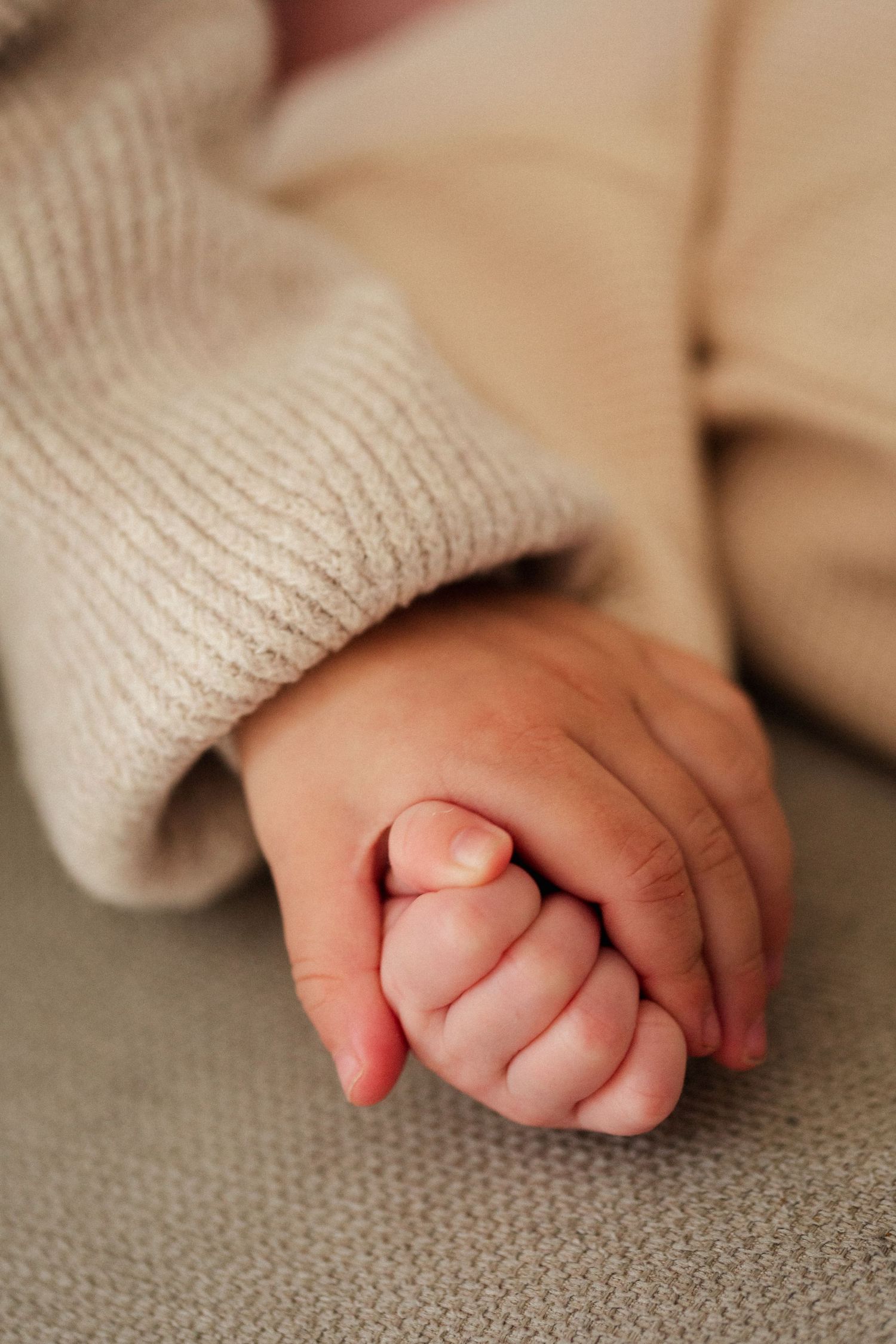 Tender moment captured as small baby hand is held by adult hand on grey surface.