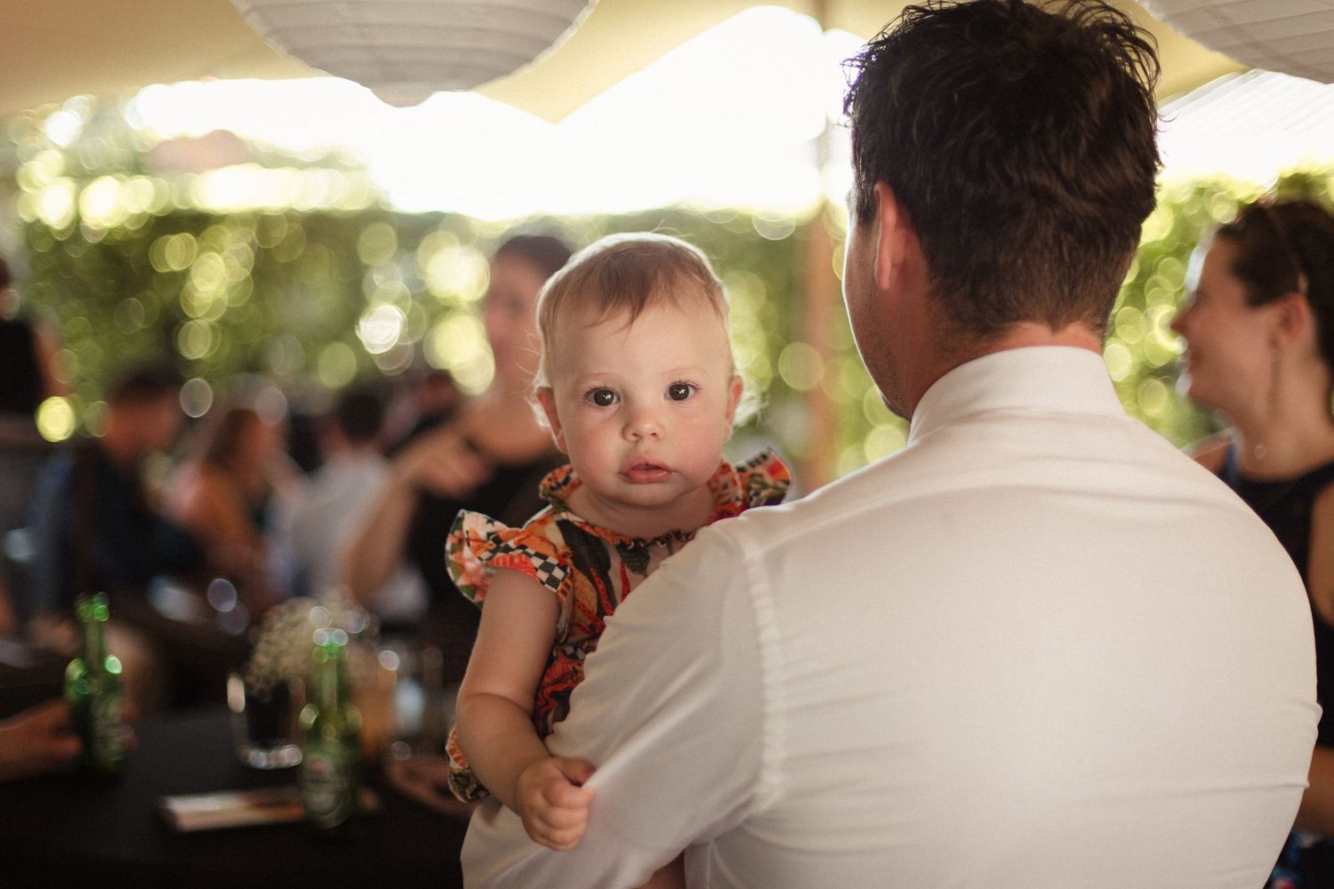 A small baby wearing a colorful dress is held at an outdoor evening reception with paper lanterns and soft lighting.