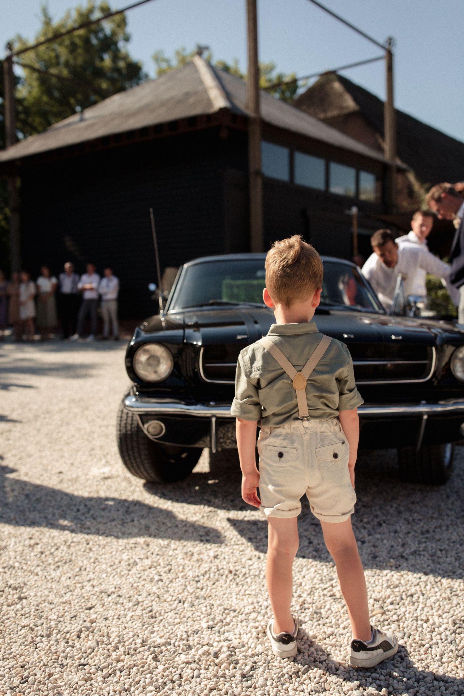 Young boy in suspenders and shorts stands in front of a classic black Ford Mustang at an outdoor event.