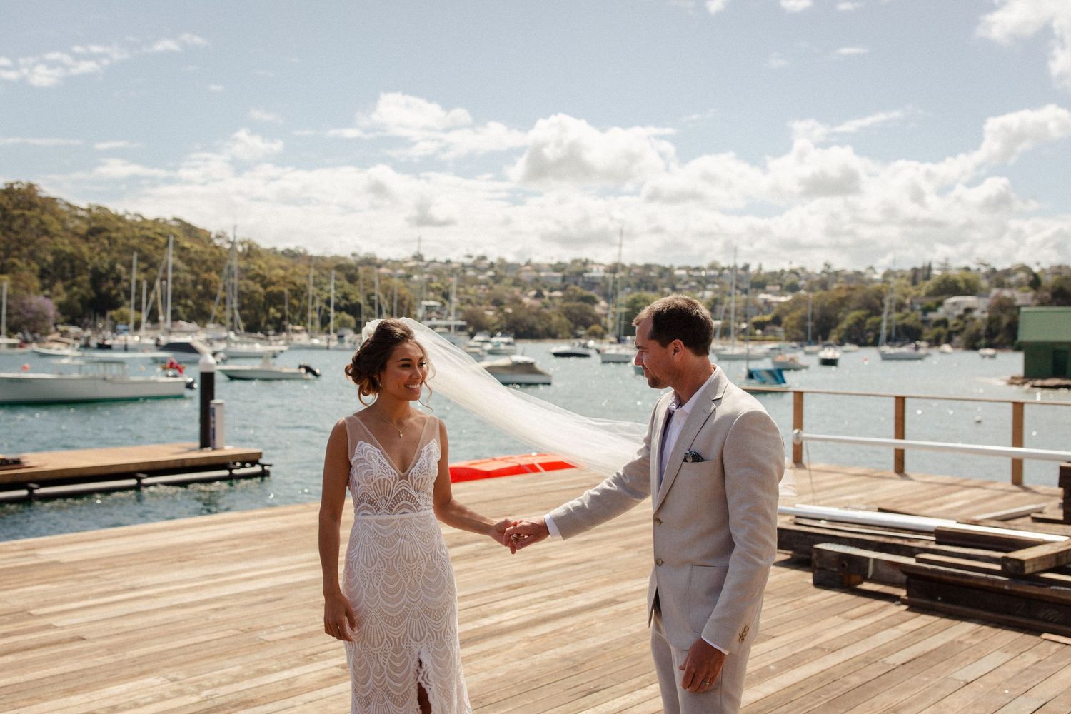 Couple in wedding attire share a moment on wooden dock with boats in harbor.