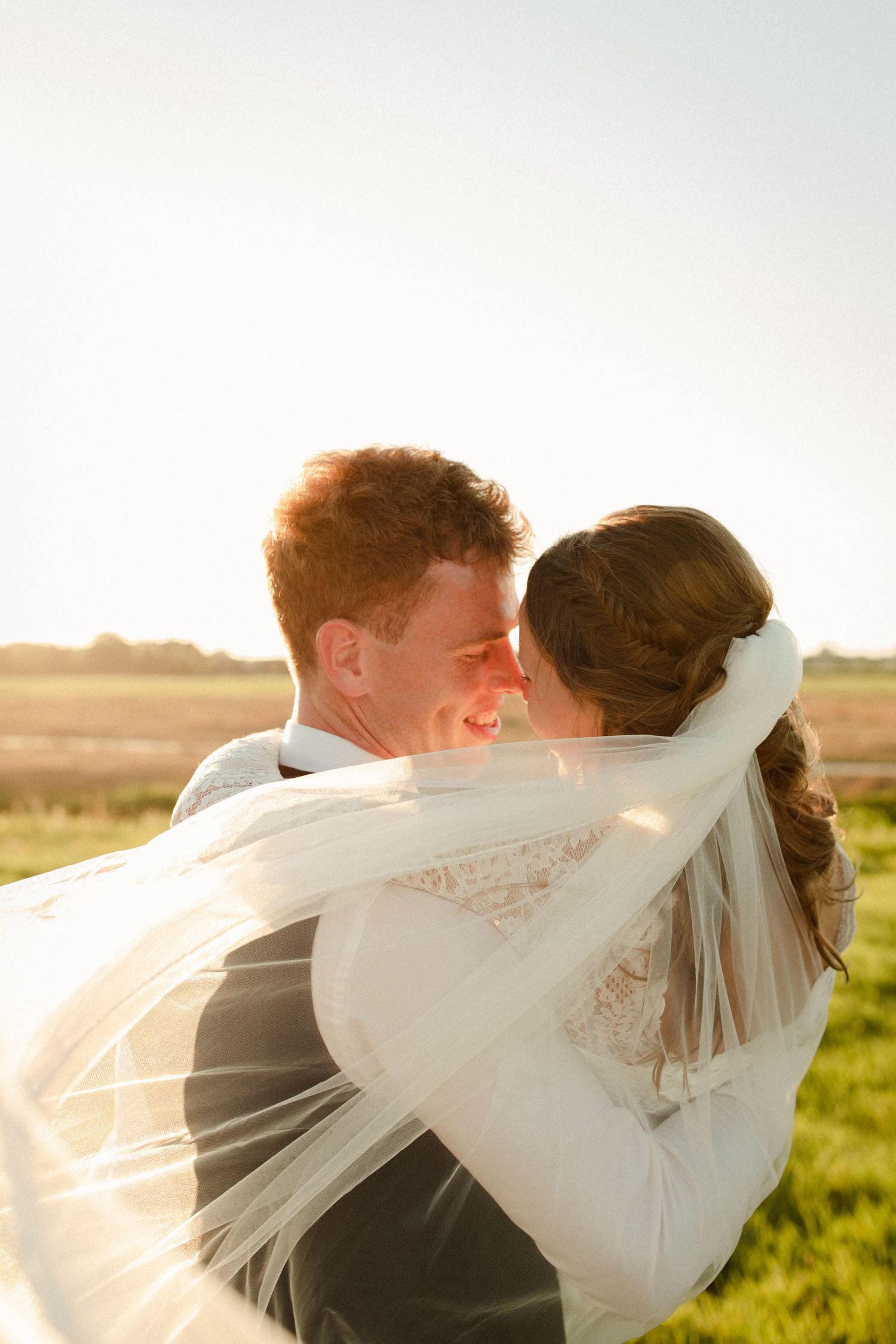 Wedding couple embraces in golden sunlight as delicate veil flows in breeze against pastoral countryside backdrop.