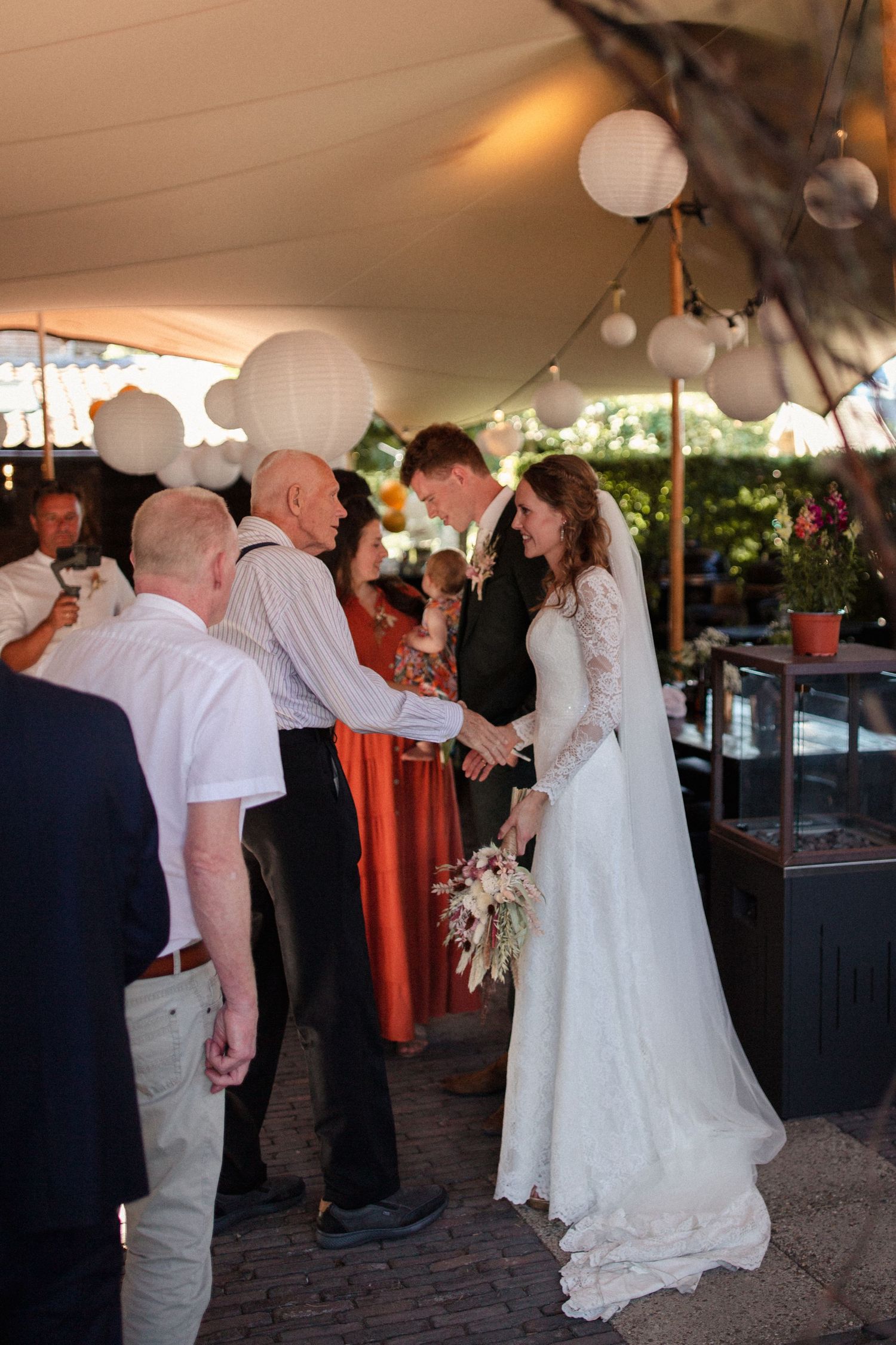 Wedding reception under a tent with glowing white lanterns as guests mingle and celebrate on a brick patio.