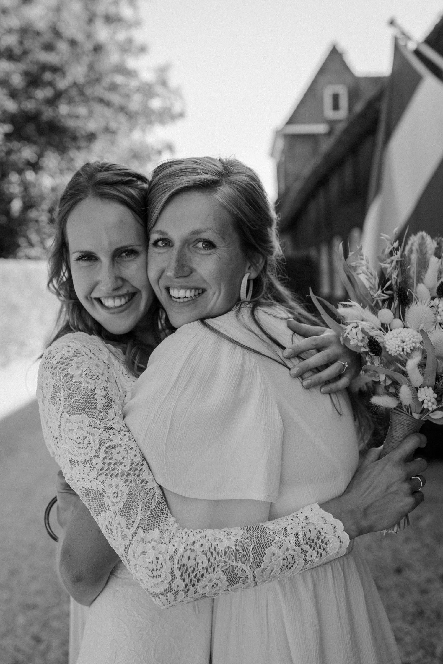 Two people in white dresses share a joyful embrace while holding a bouquet outdoors.