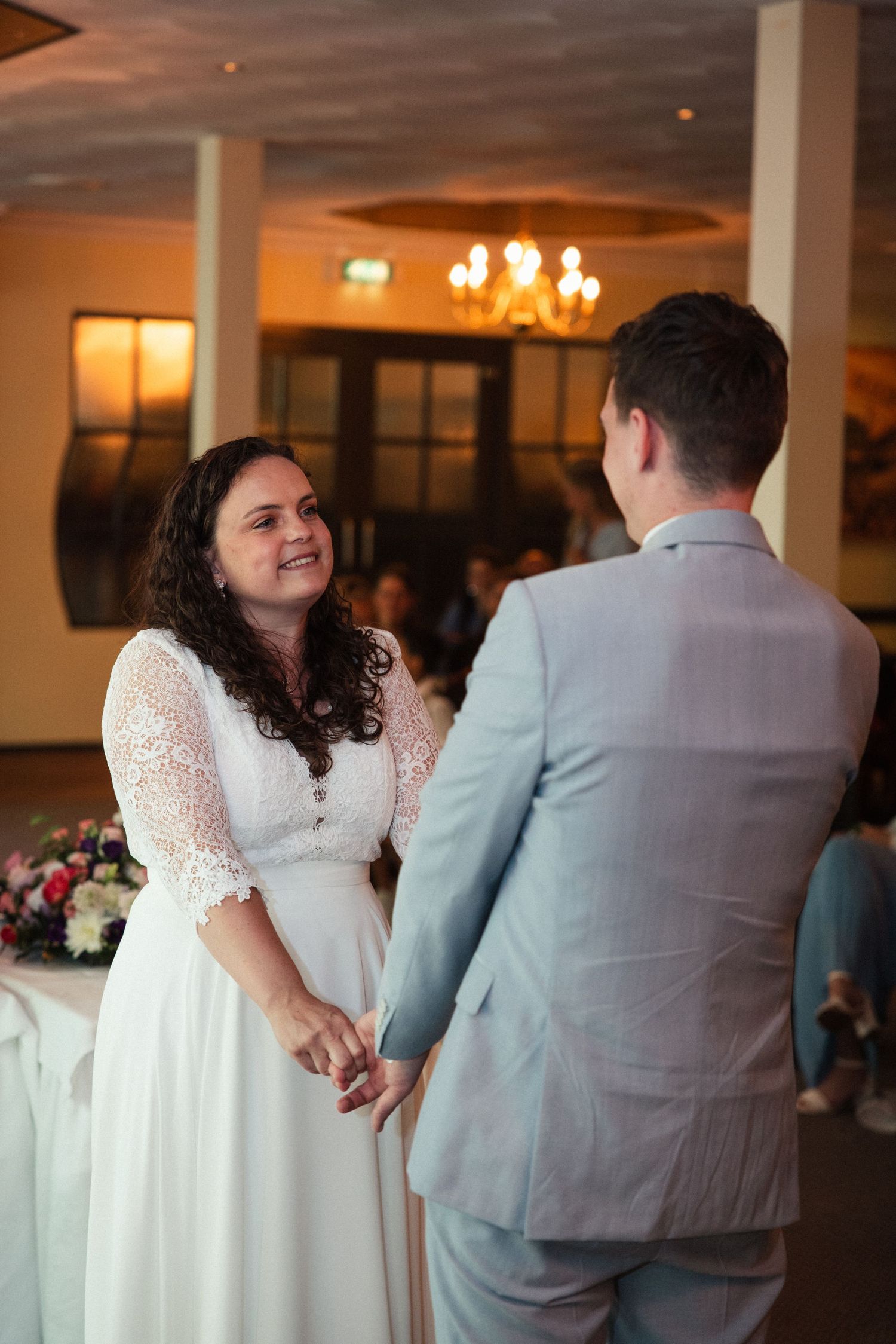A lace-gown wedding couple shares an intimate moment while dancing under a warm chandelier in an elegant ballroom.