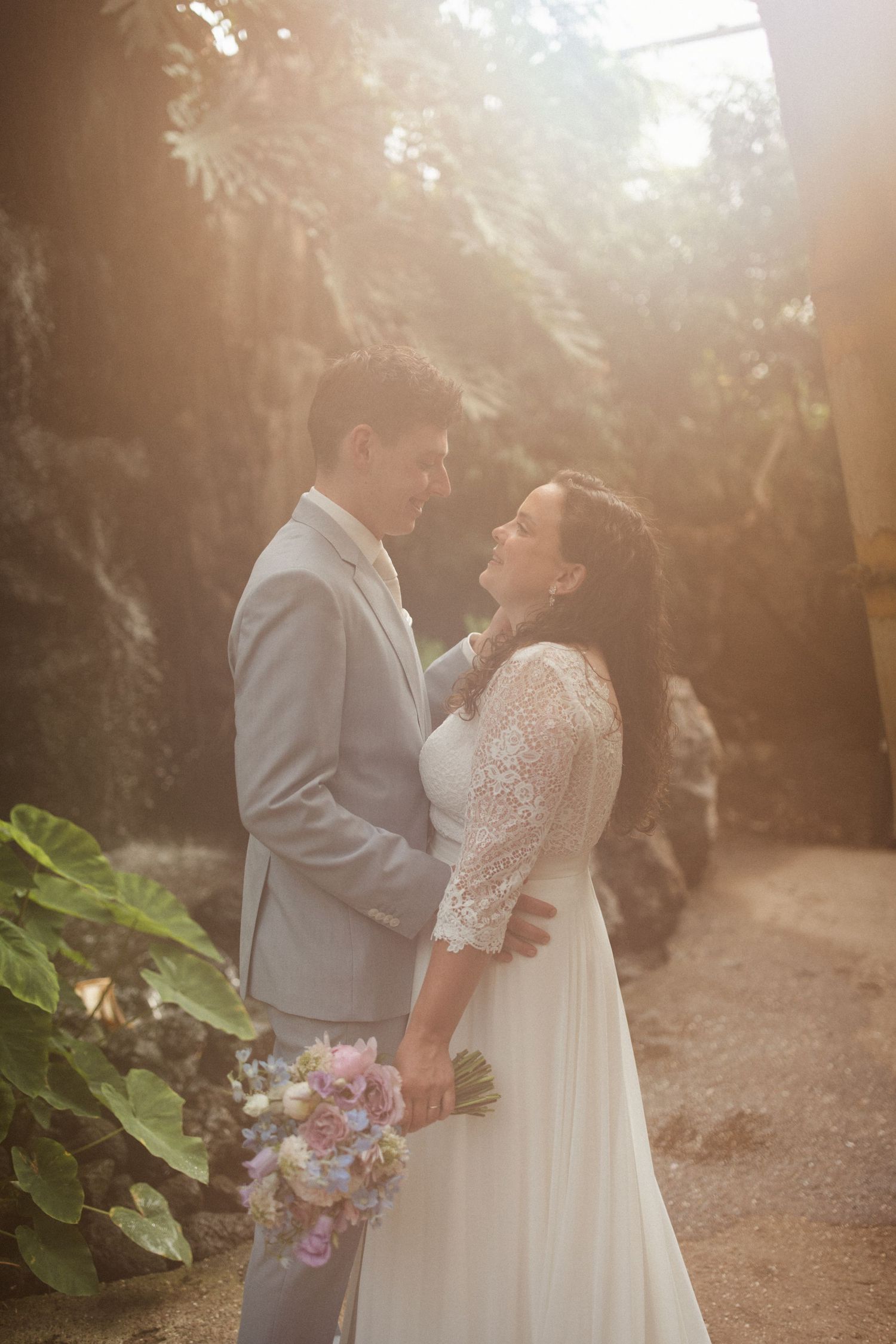 A couple shares romantic moments in golden sunlight surrounded by lush greenery while holding a floral bouquet.