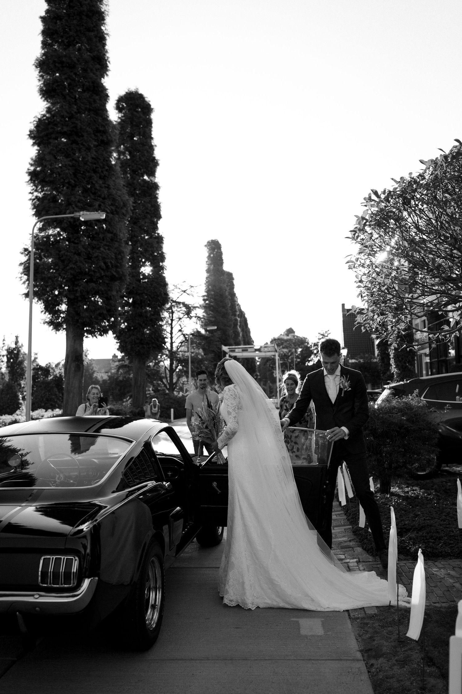 Black and white photo of newlyweds next to a vintage Ford Mustang after their wedding ceremony.