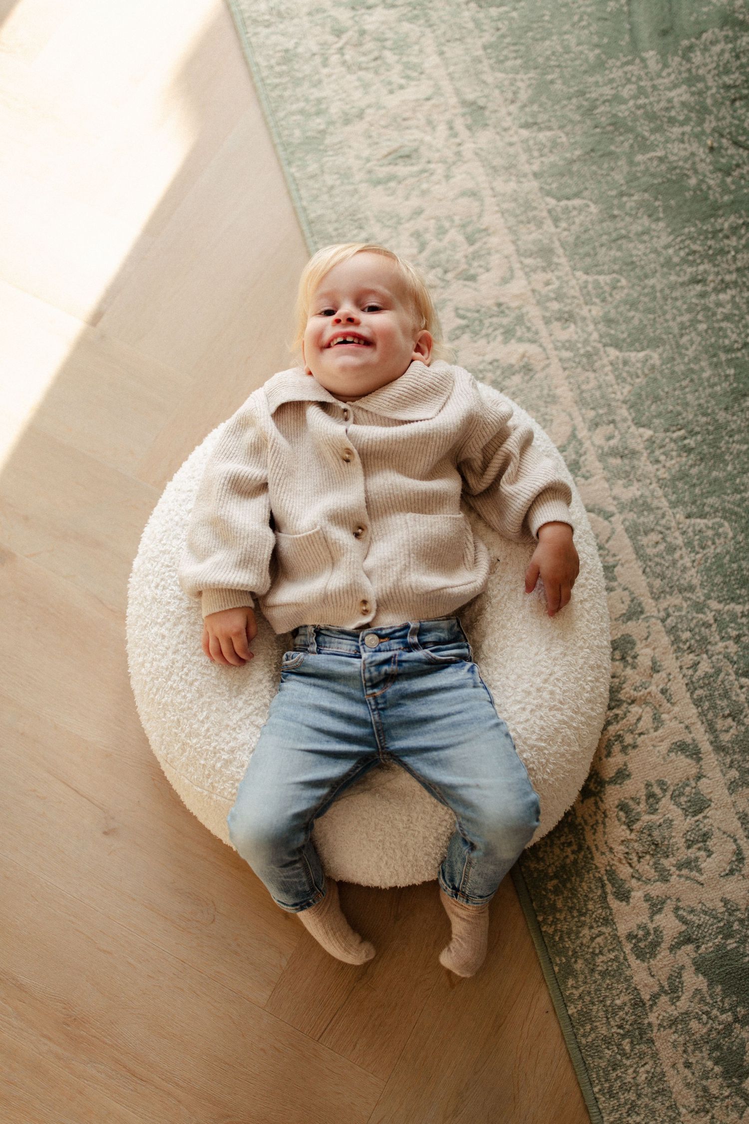 Smiling baby wearing beige cardigan and jeans sitting on round white cushion.