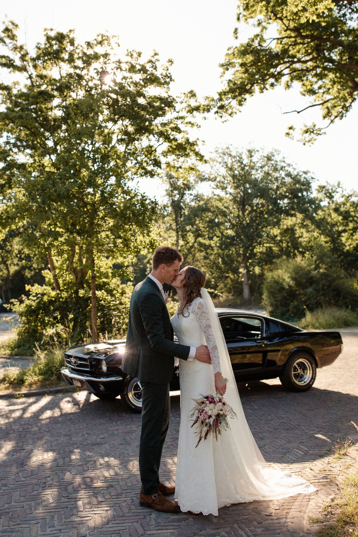 A classic black Mustang car parked on a brick road with sun filtering through trees creates a romantic backdrop.