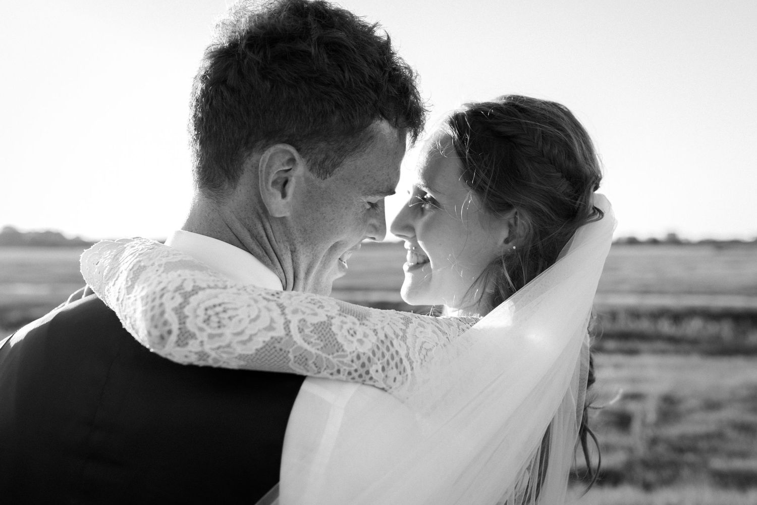 Black and white intimate wedding portrait showing couple embracing in rural setting with long veil and lace details.