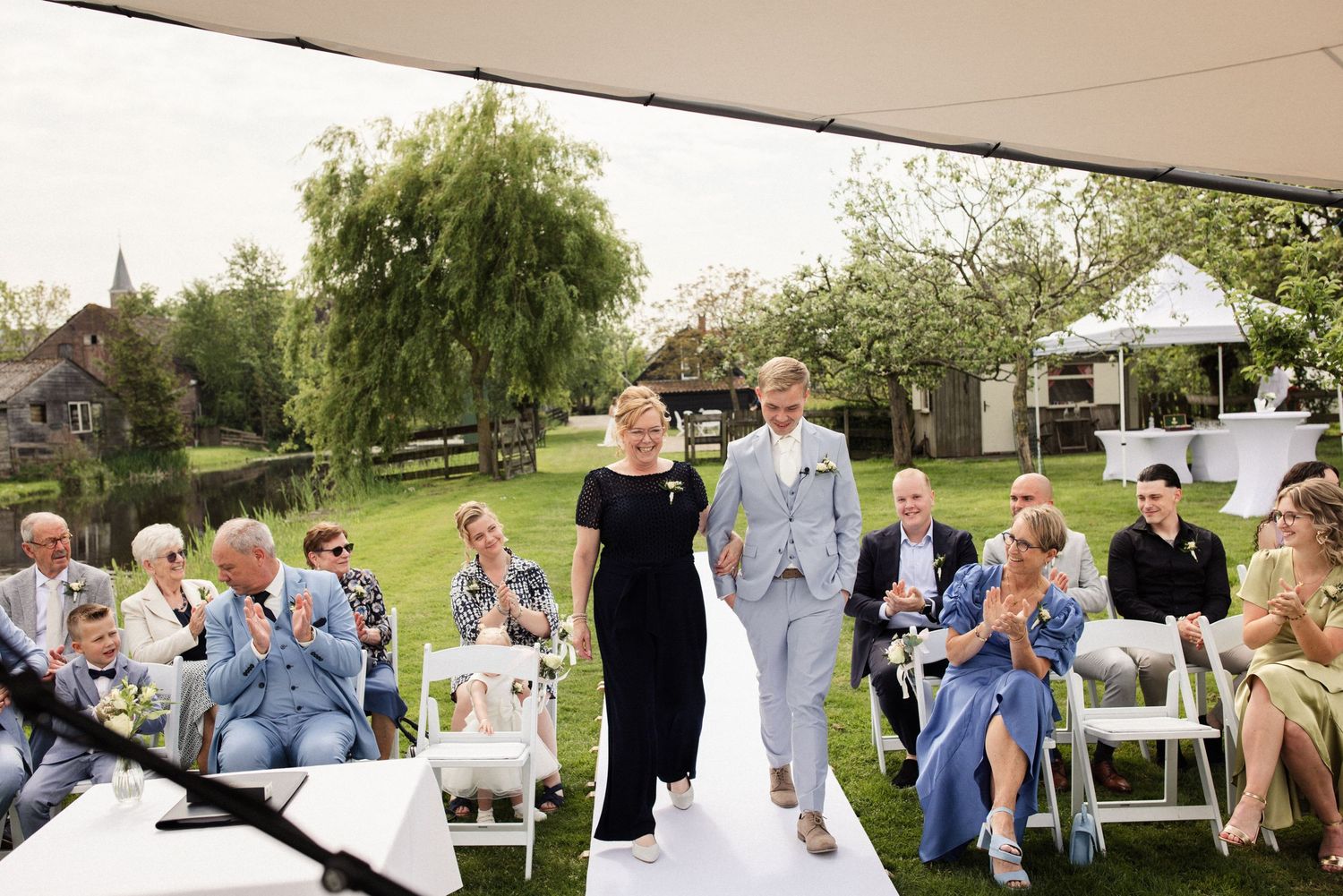 An elegant outdoor wedding ceremony in a garden setting with guests seated on white chairs and a white aisle runner.