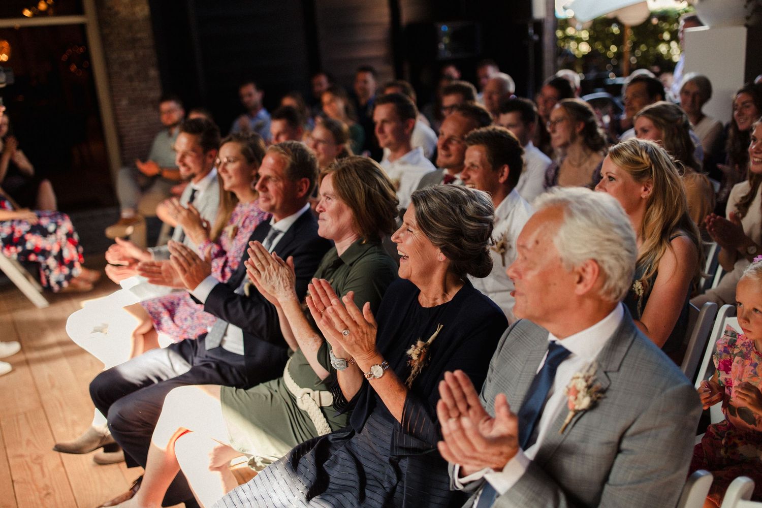 Wedding guests applaud during an indoor ceremony while seated in rows with warm lighting.