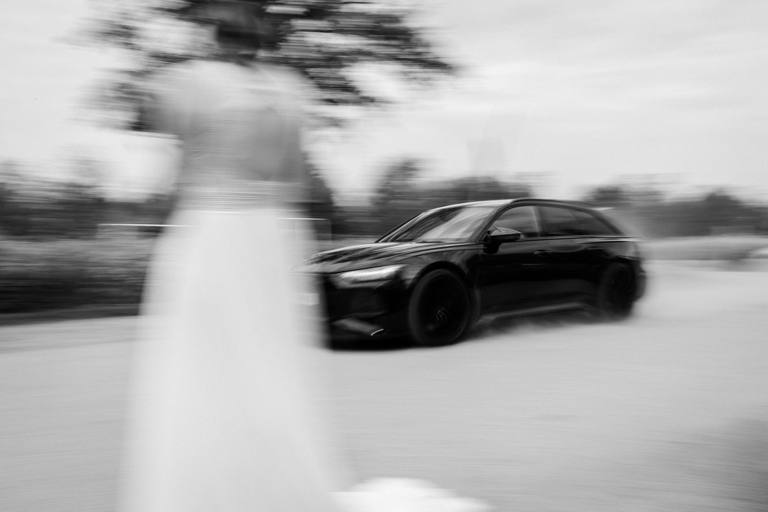 Blurred motion black luxury car passes by white figure in dreamy black and white photography.