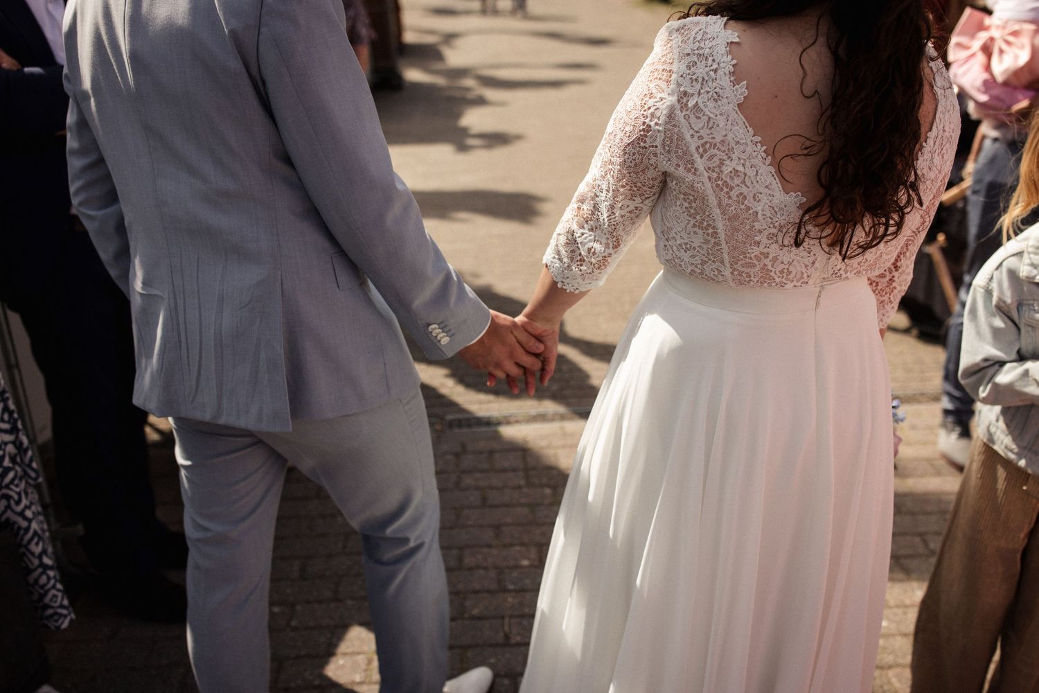 A couple in wedding attire holds hands while walking on a sunny brick pathway.
