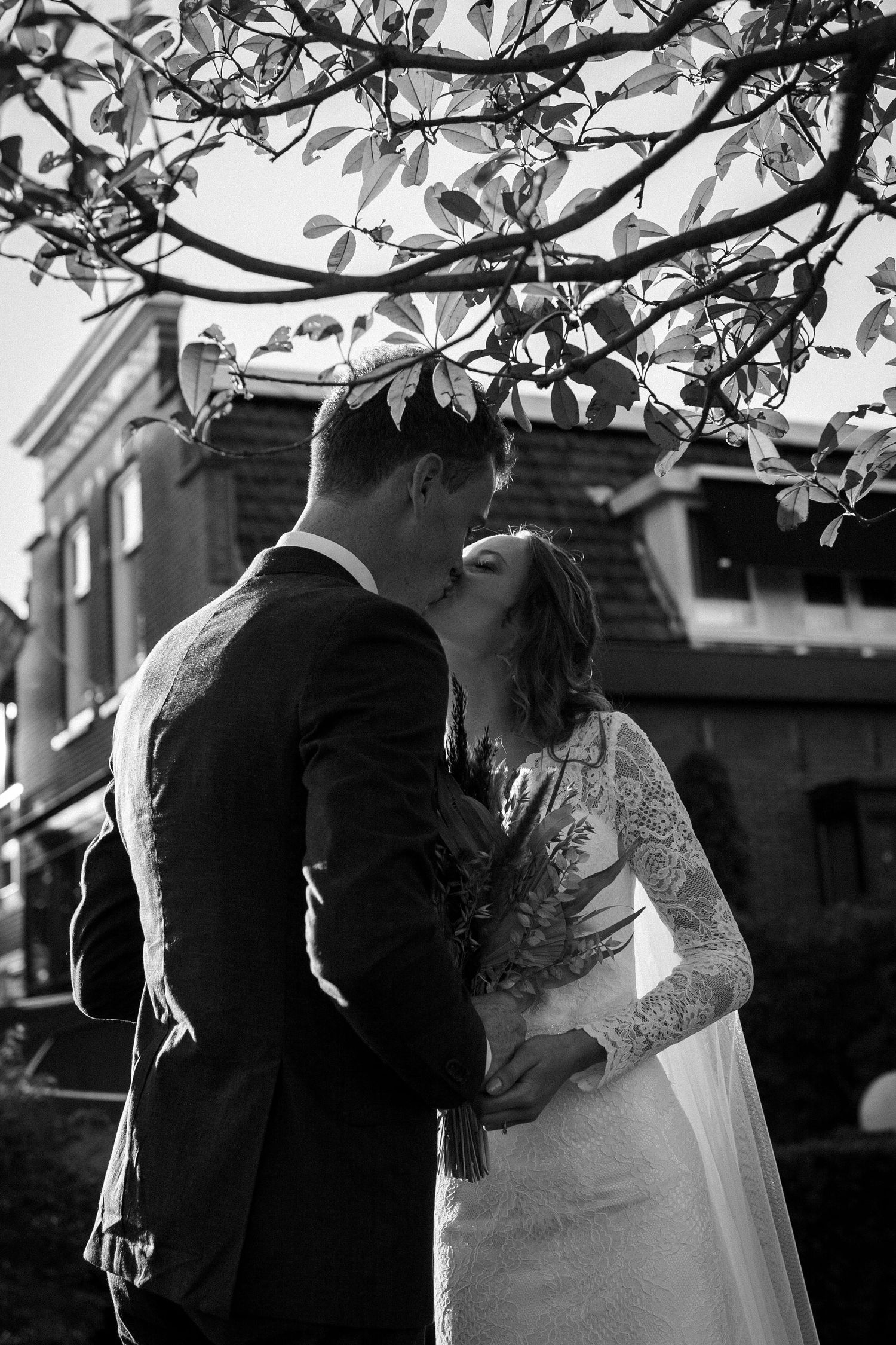 A romantic black and white wedding photograph showing a couple kissing under tree branches in front of a building.