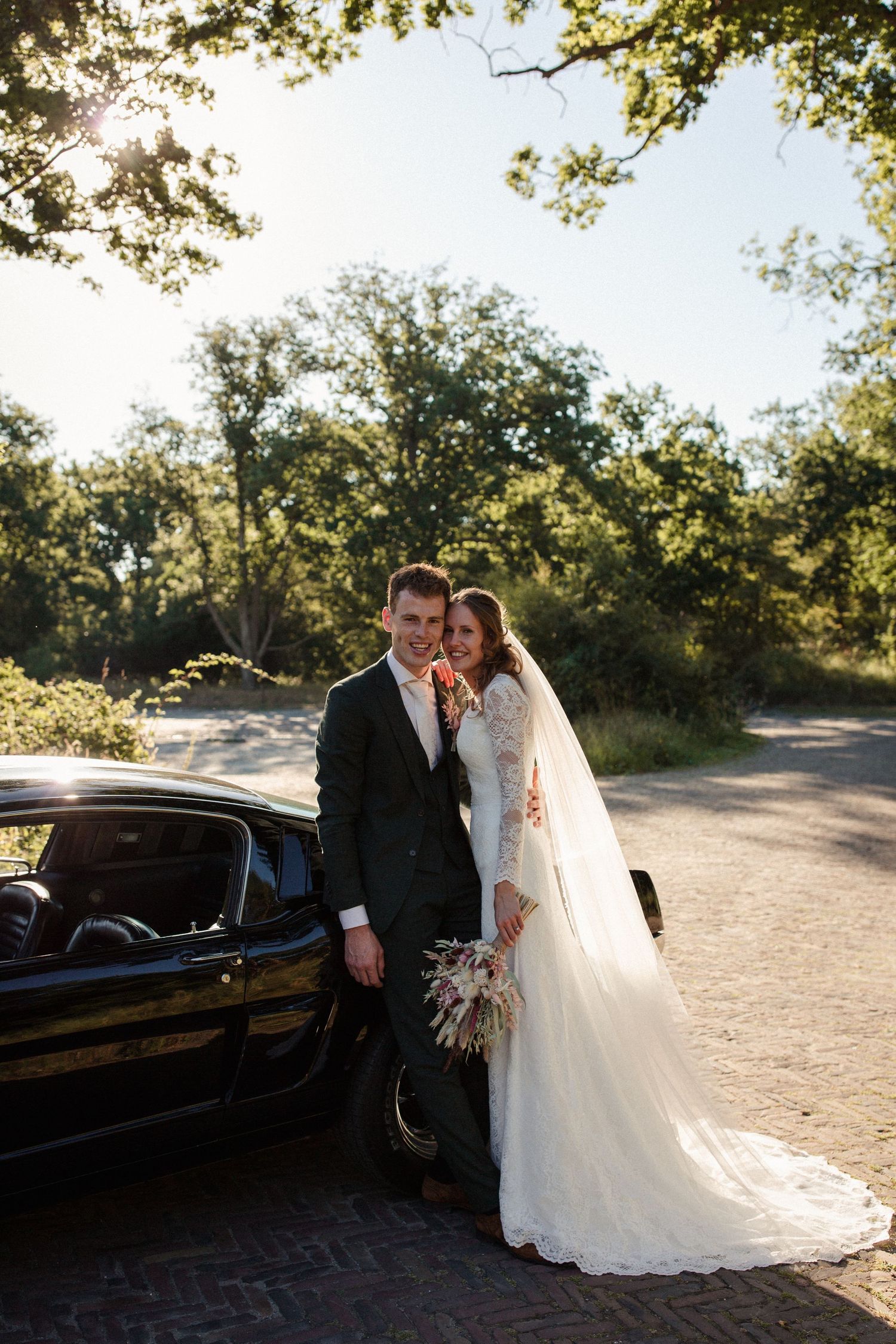 A wedding couple stands next to a classic black car on a sunny day with trees in the background.