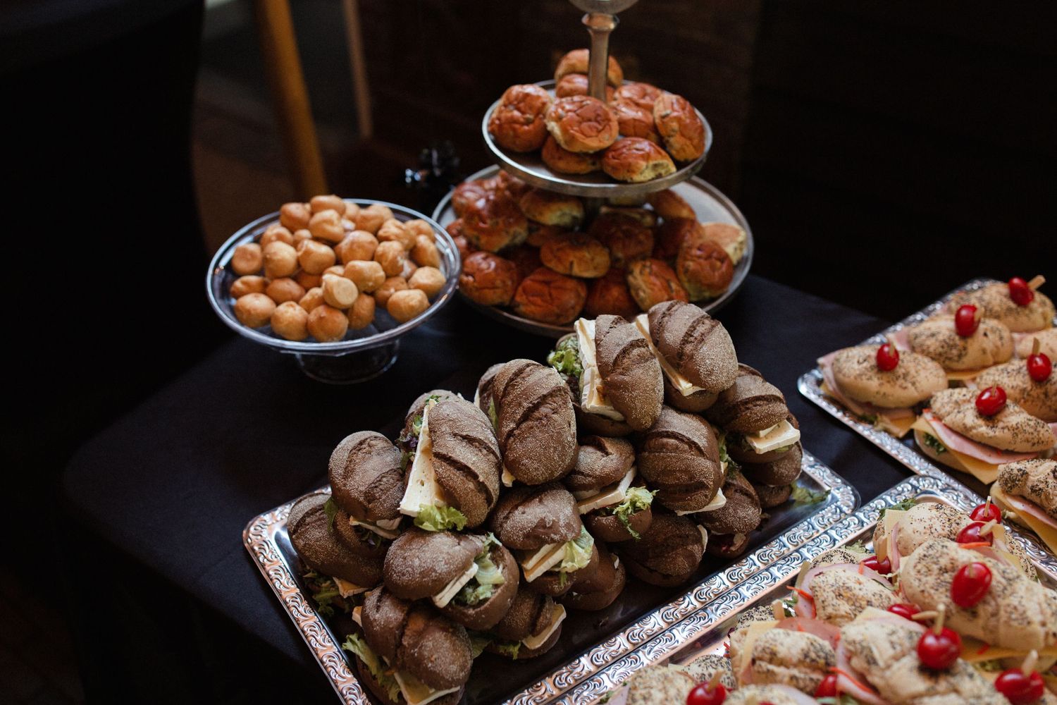 An elegant party spread featuring stacked sandwiches, pastries, and desserts arranged on a dark table surface.