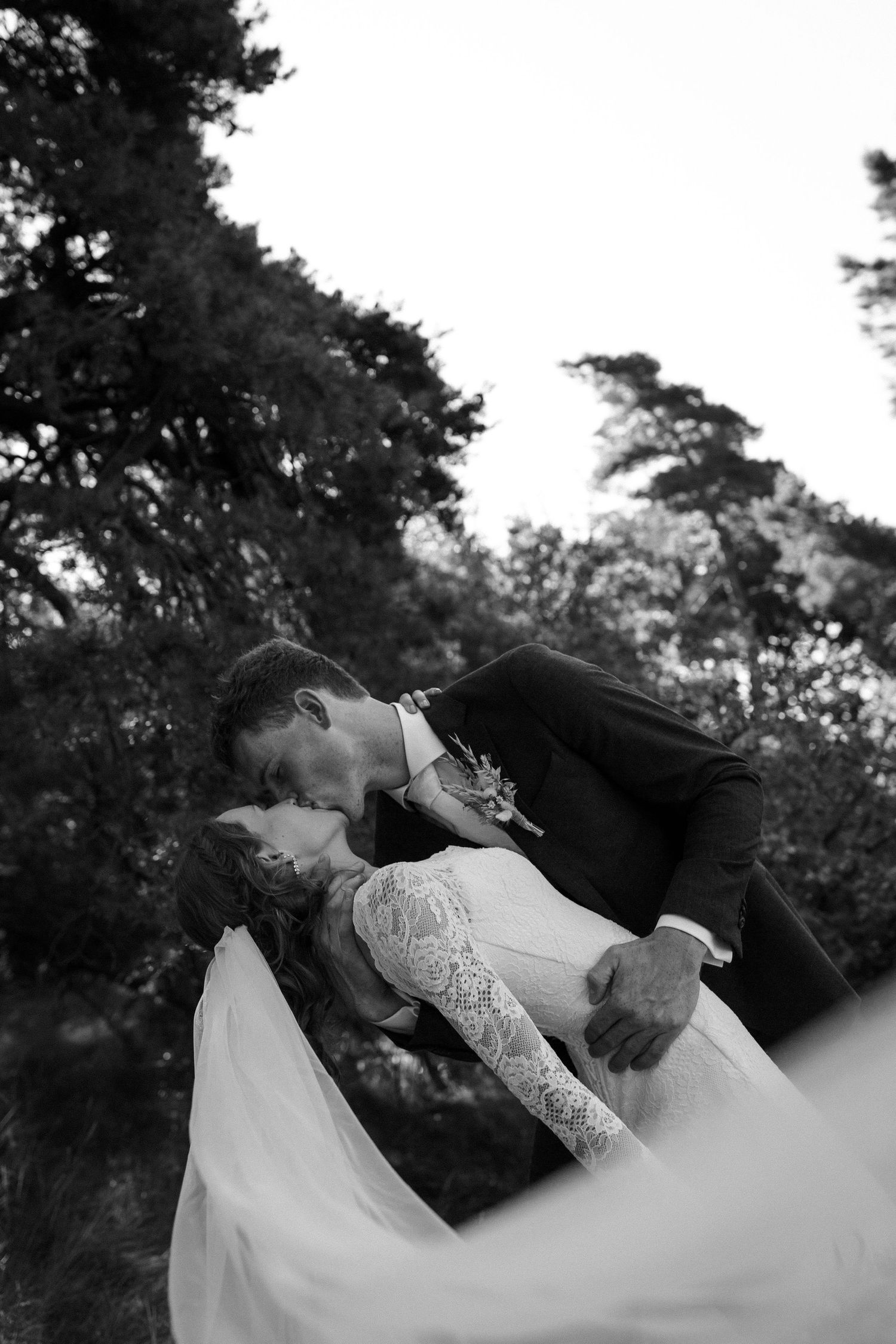 A black and white romantic wedding photograph showing a couple embracing in a dramatic dip pose among trees.