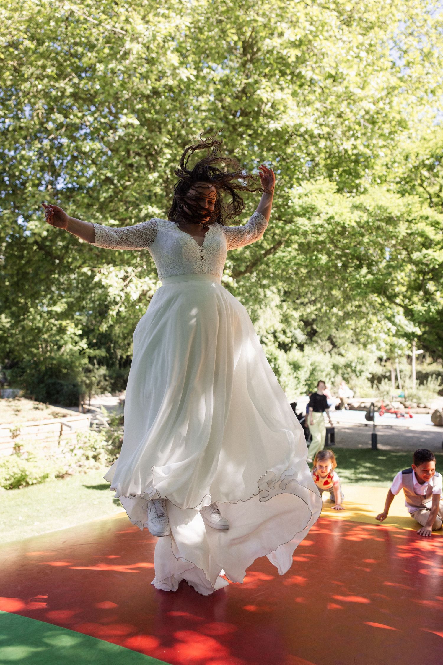 A person in a flowing white wedding dress joyfully leaps on a colorful outdoor dance floor surrounded by greenery.