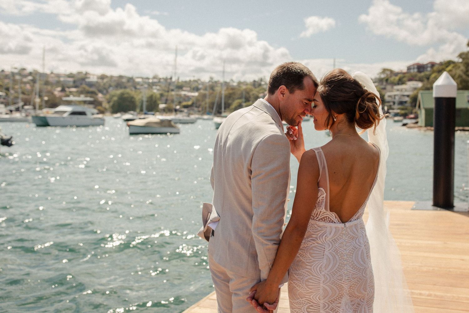 Newlyweds share an intimate moment on a sunny dock with boats and water behind them.