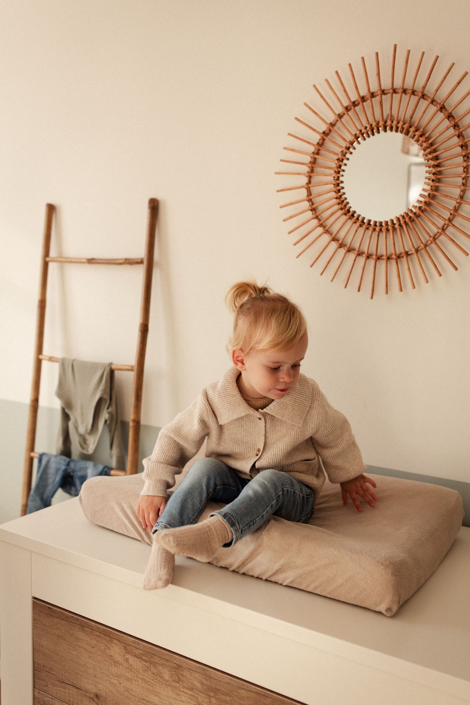 Child in beige cardigan sits on cushion near decorative sunburst mirror and ladder.