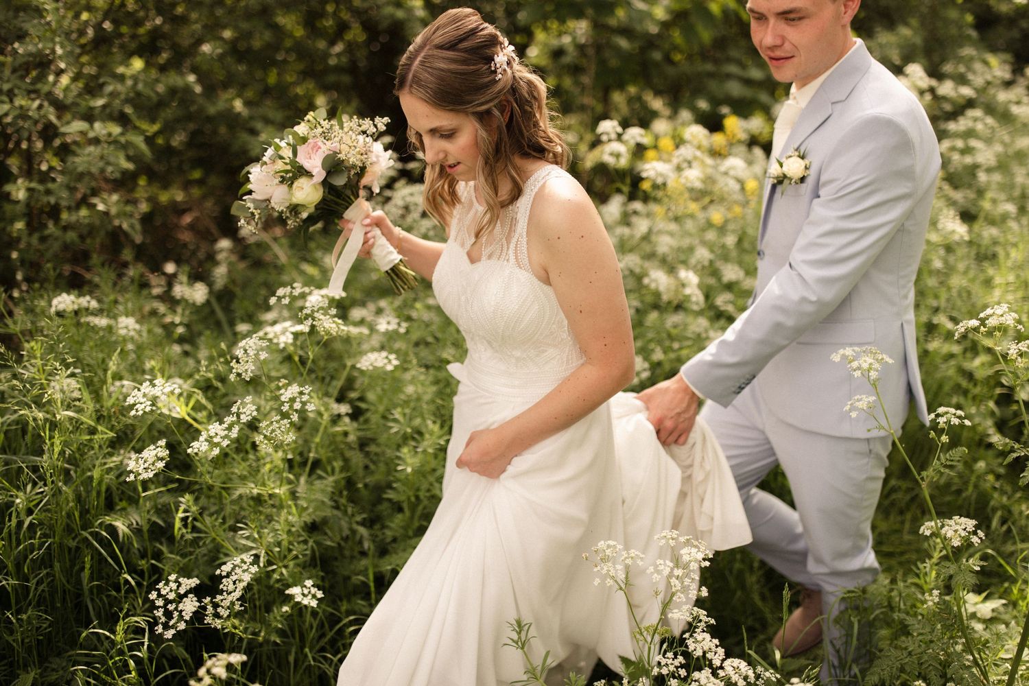 Newlyweds in white dress and light suit share a tender moment in a garden with summer flowers.