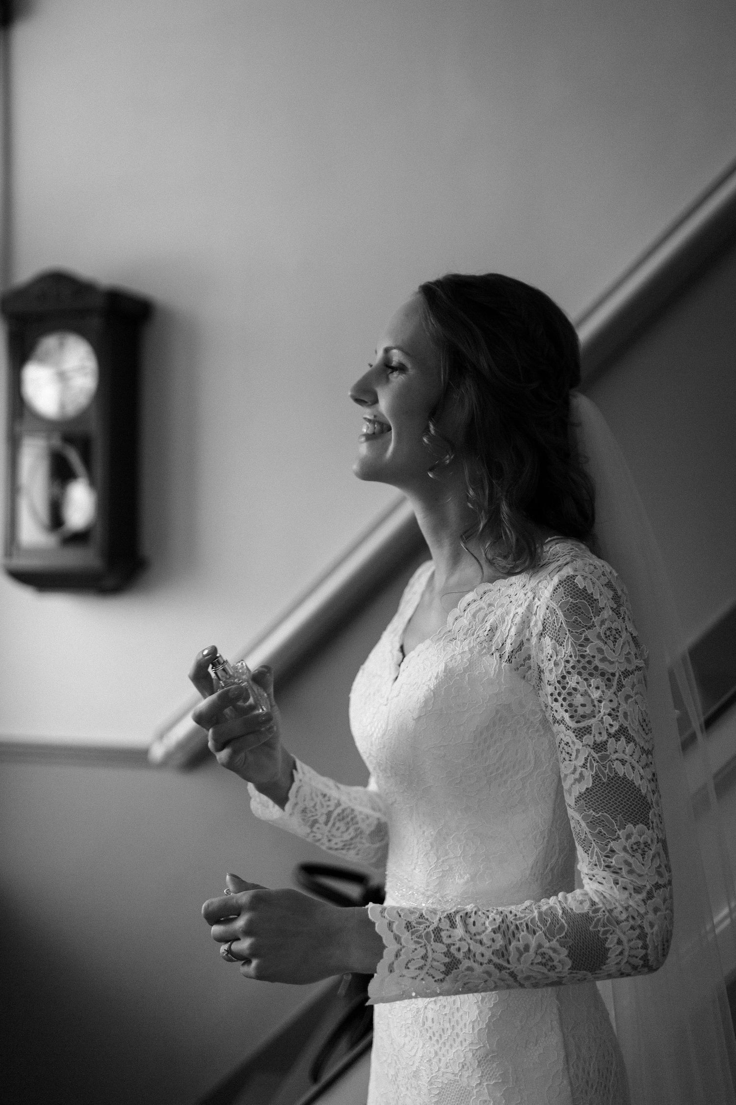 A black and white photograph of a bride in a lace long-sleeved wedding dress laughing joyfully beside a vintage wall clock.