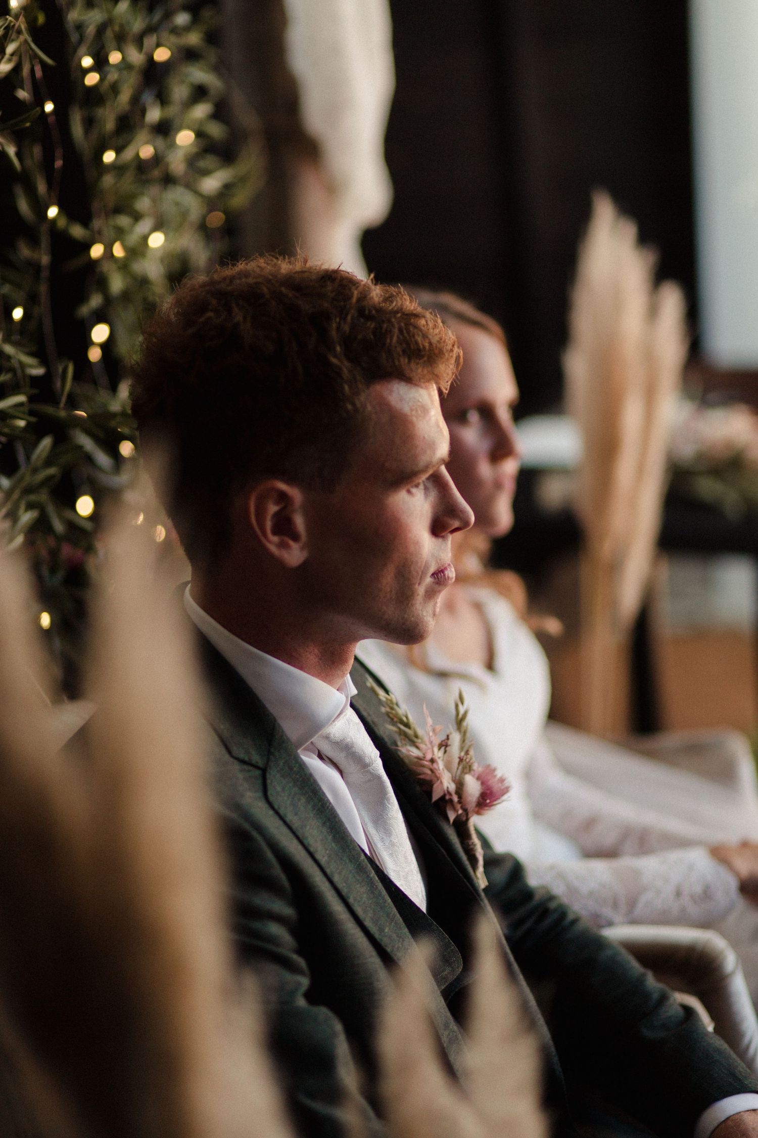 A couple sits in formal attire at a wedding ceremony with a decorated Christmas tree and pampas grass in the background.