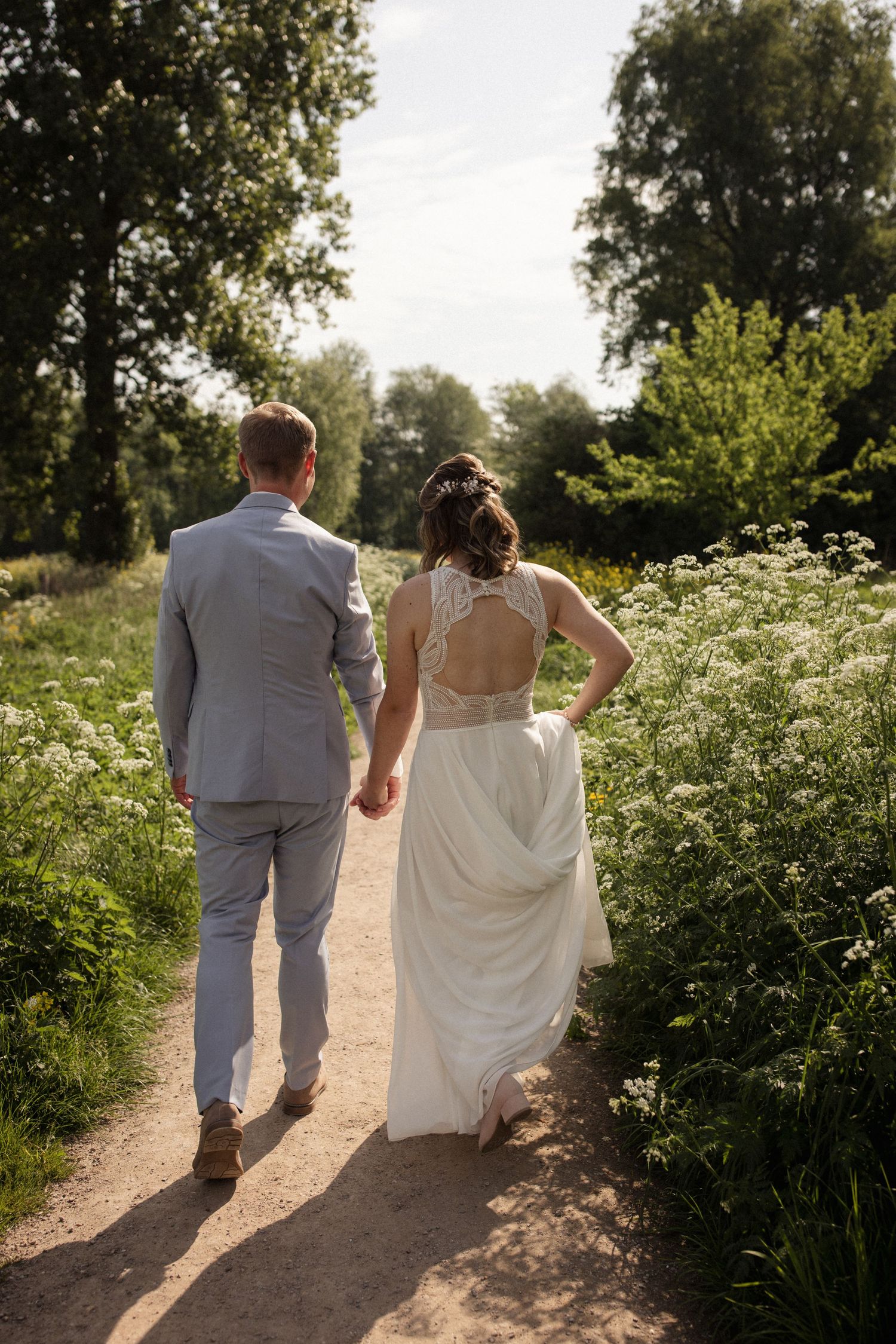 A couple in wedding attire holds hands while walking down a wildflower-lined path in dappled sunlight.