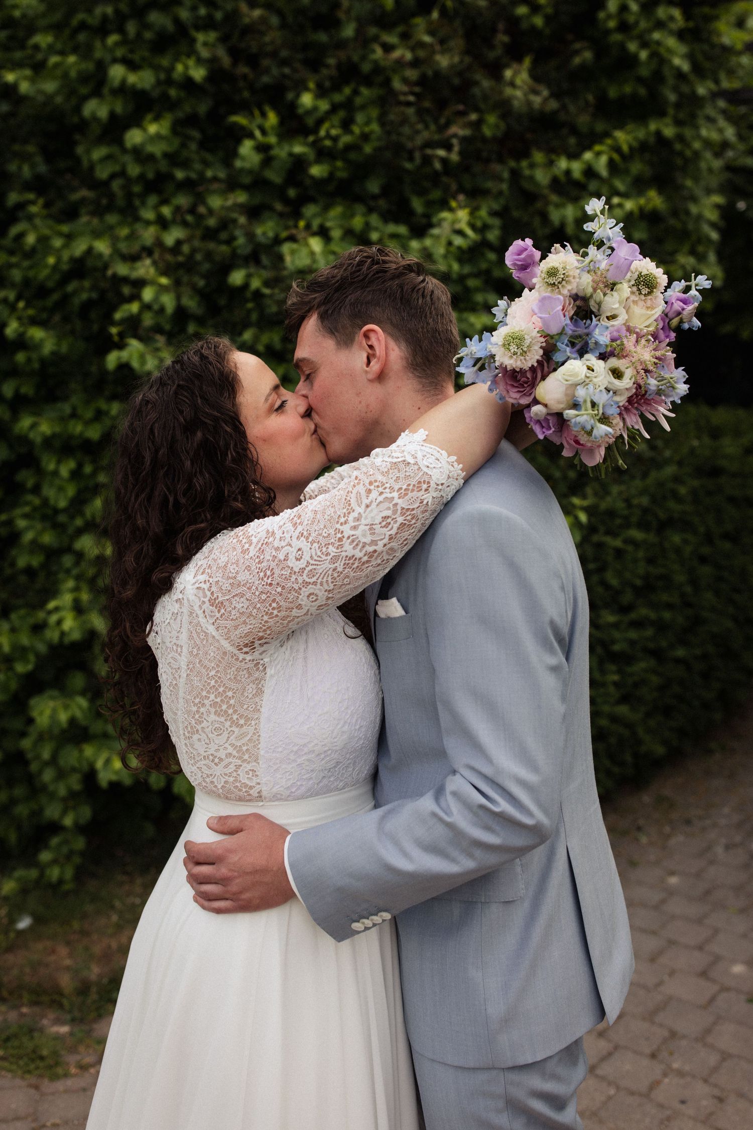 Newlyweds share romantic kiss while bride holds beautiful pastel bouquet against garden backdrop.
