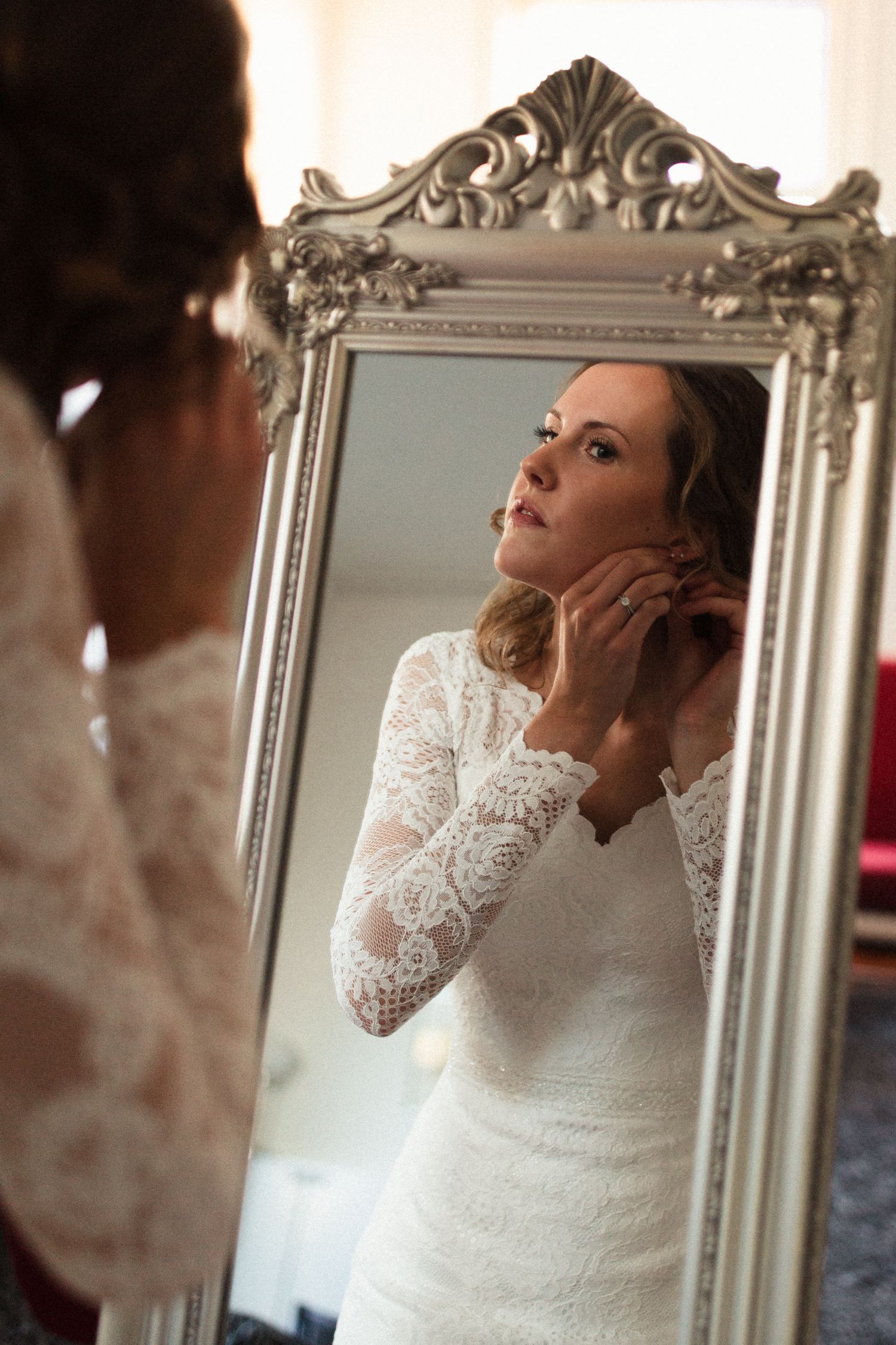 A reflection in an ornate silver mirror shows a person in a white lace long-sleeve wedding dress adjusting jewelry.