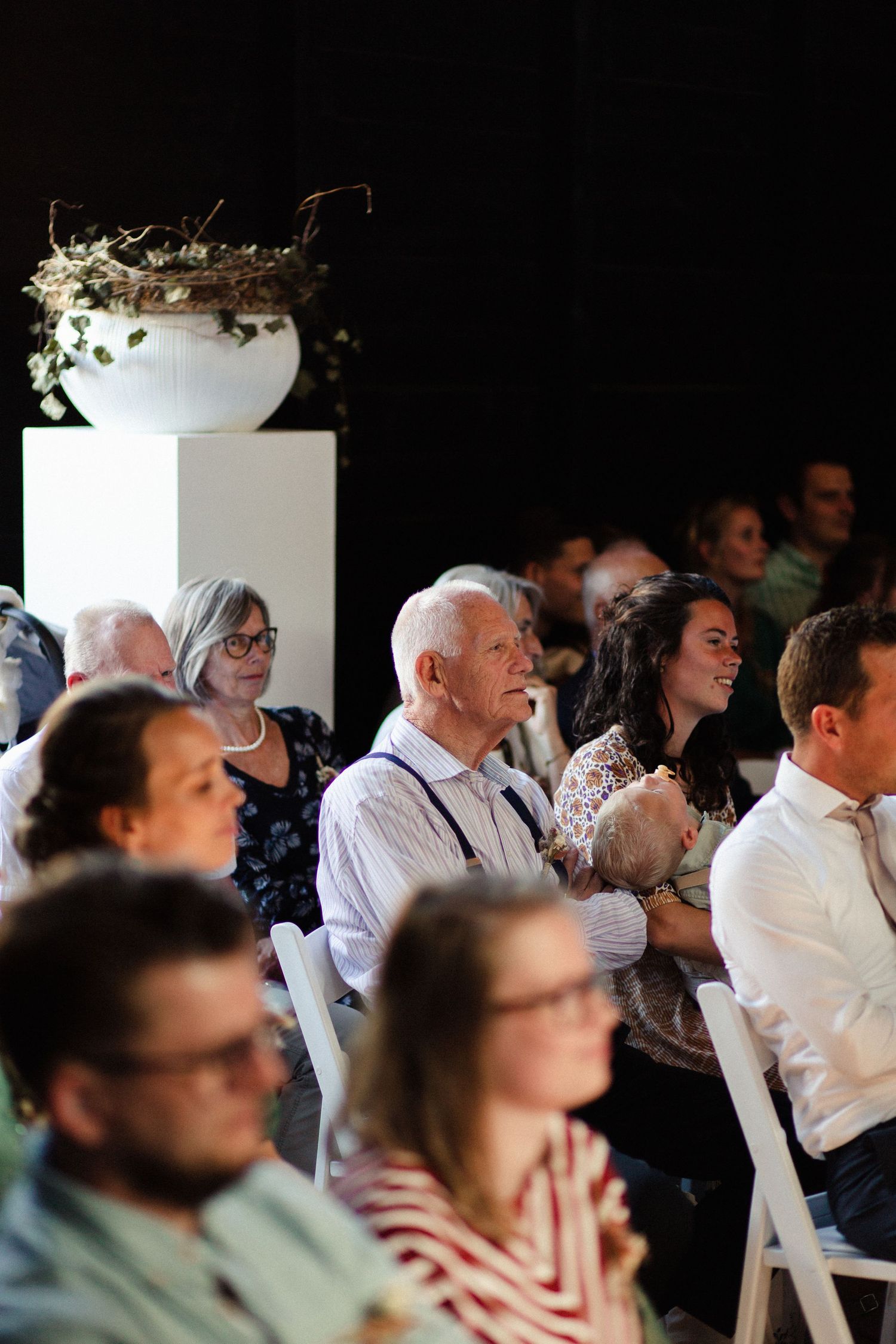 Wedding guests sit attentively in white chairs during an indoor ceremony with decorative floral arrangements.