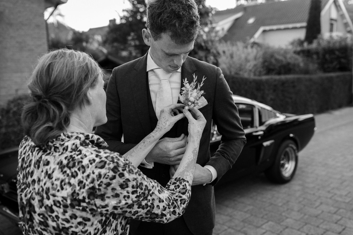 A black and white photo shows someone pinning a boutonniere on a suit jacket while standing near a car.