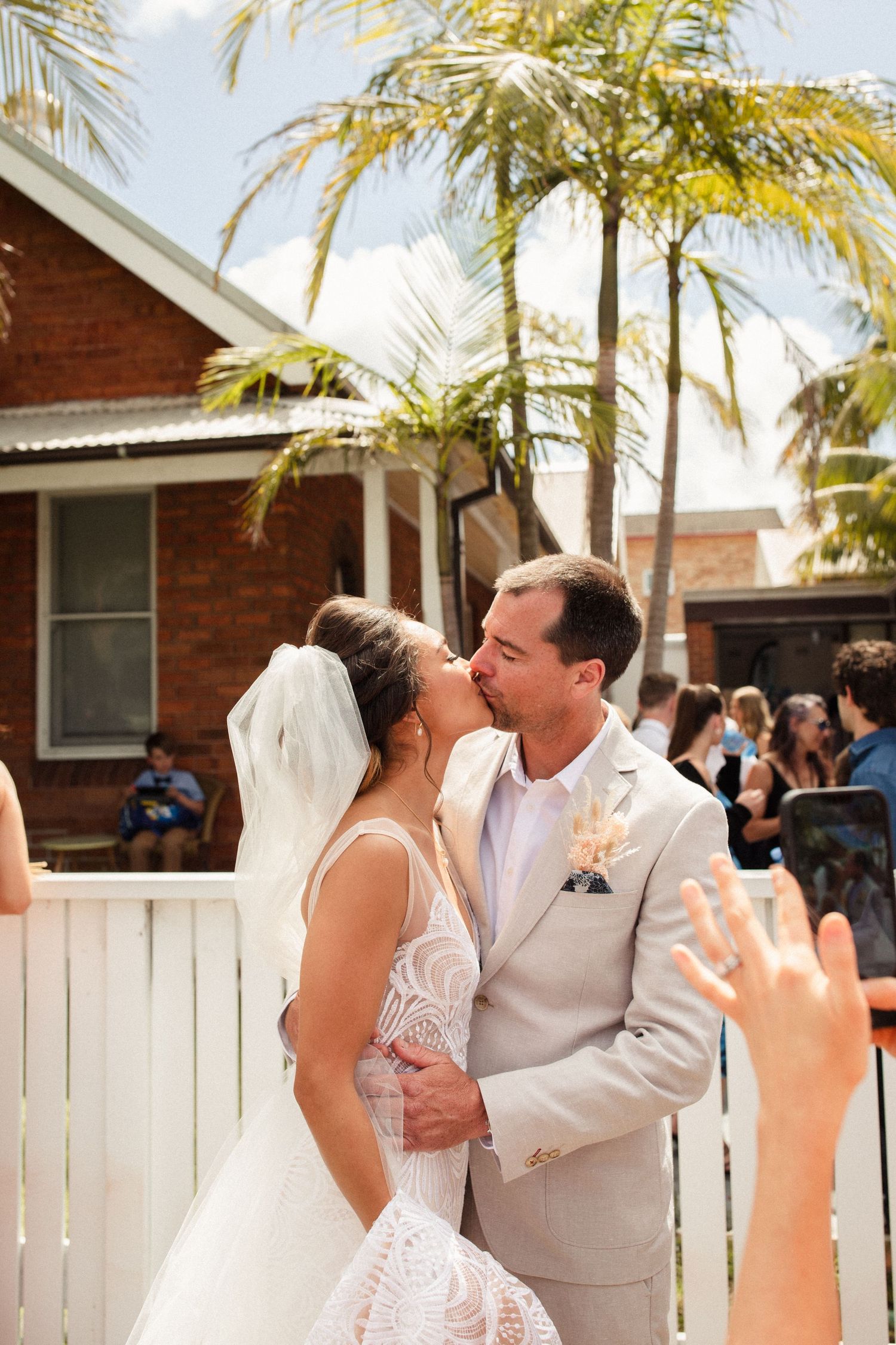 Newlyweds share their first kiss under swaying palm trees outside a brick building.
