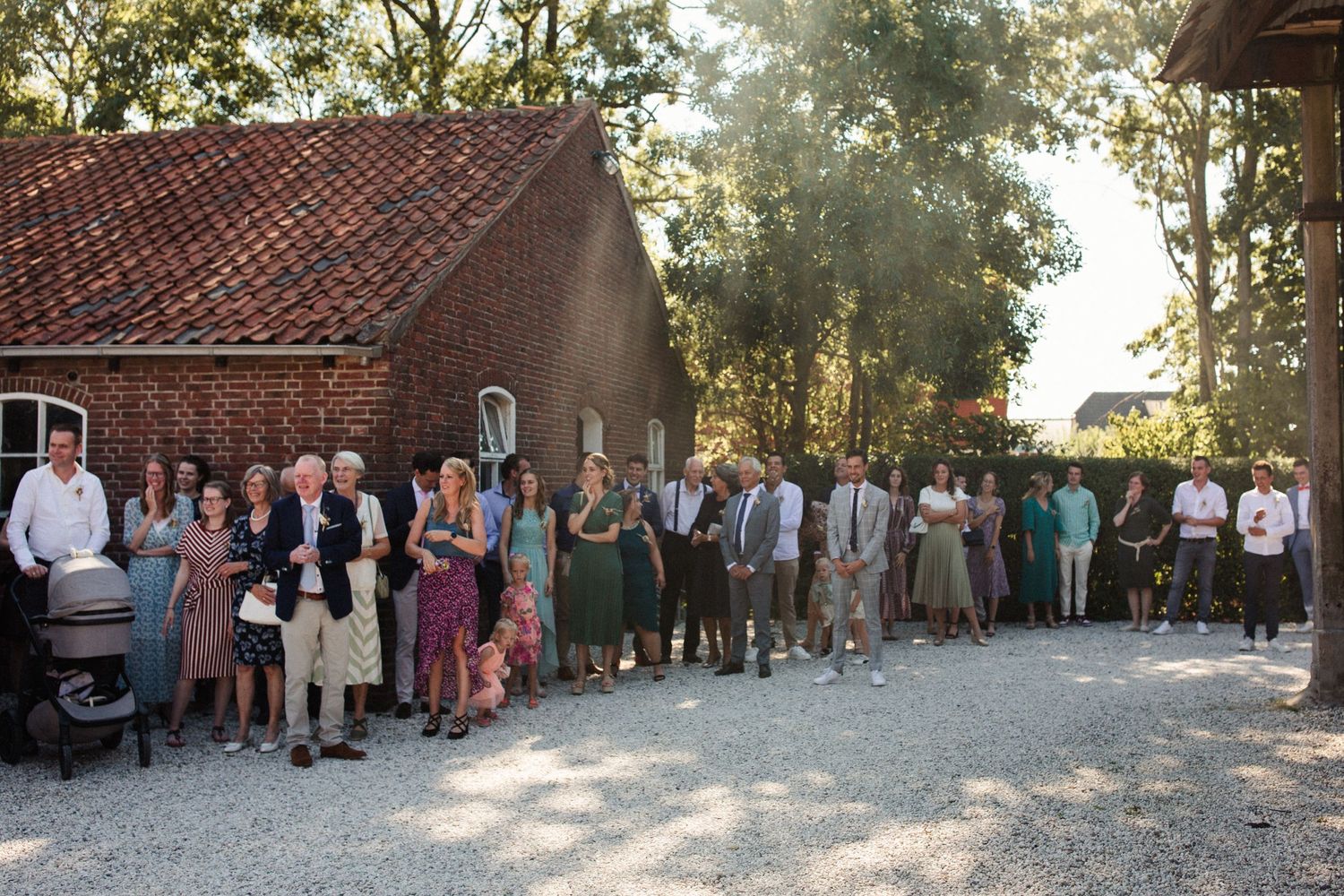 A group of wedding guests stands in line outside a rustic brick building on a sunny afternoon.