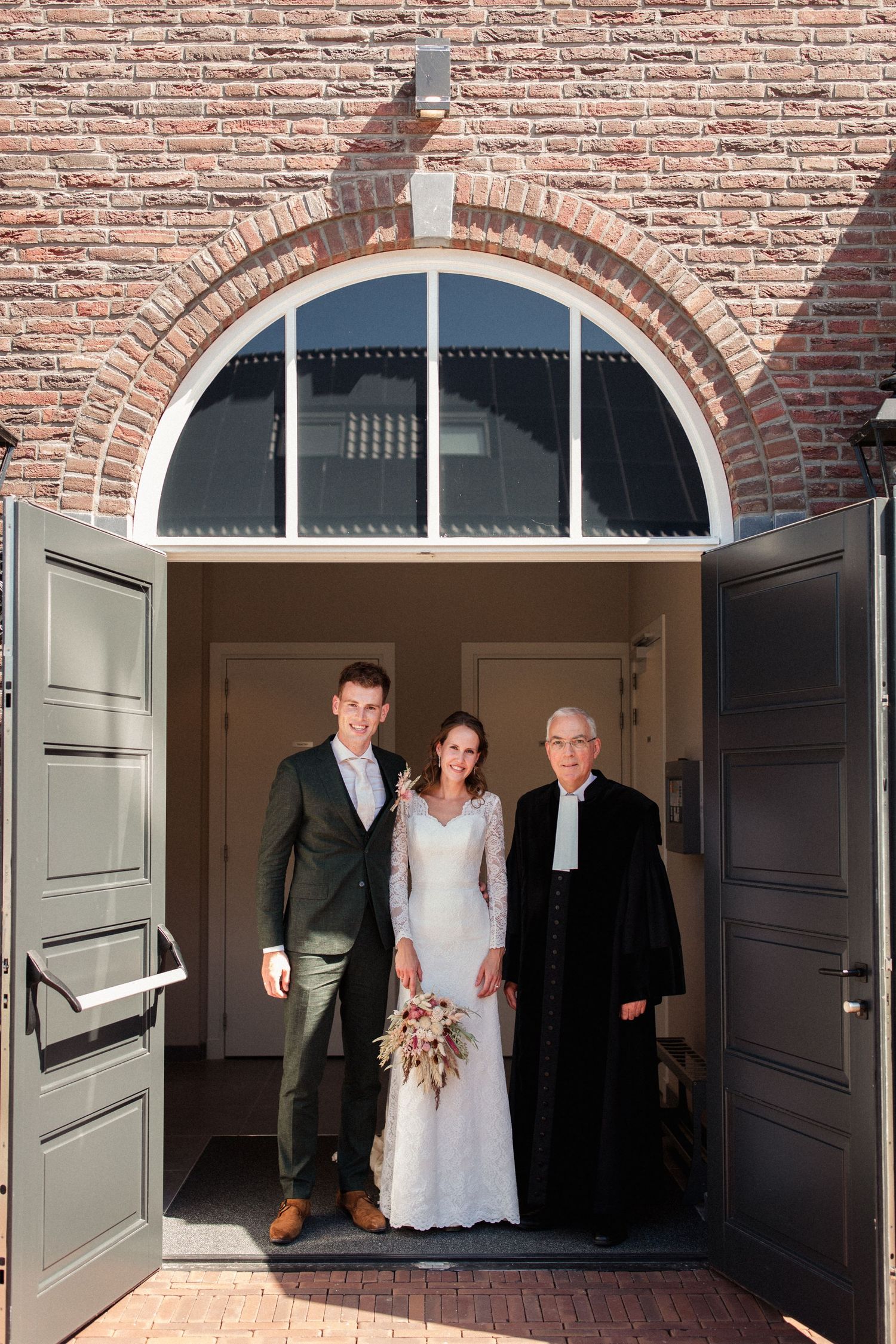 Three people stand in a brick church doorway under an arched entrance during a wedding ceremony.