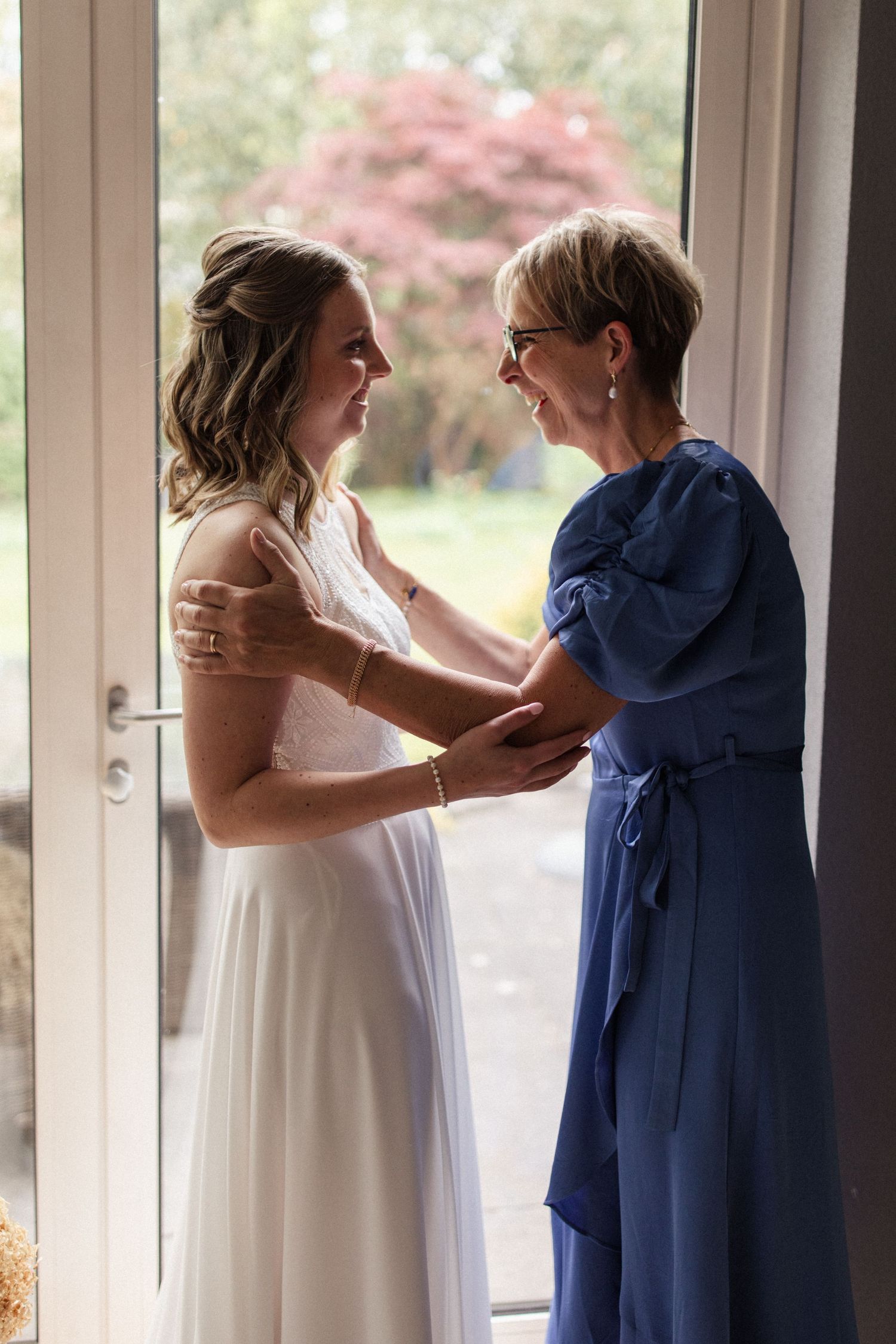 Two people share an intimate moment in formal attire by a window with pink cherry blossoms visible outside.