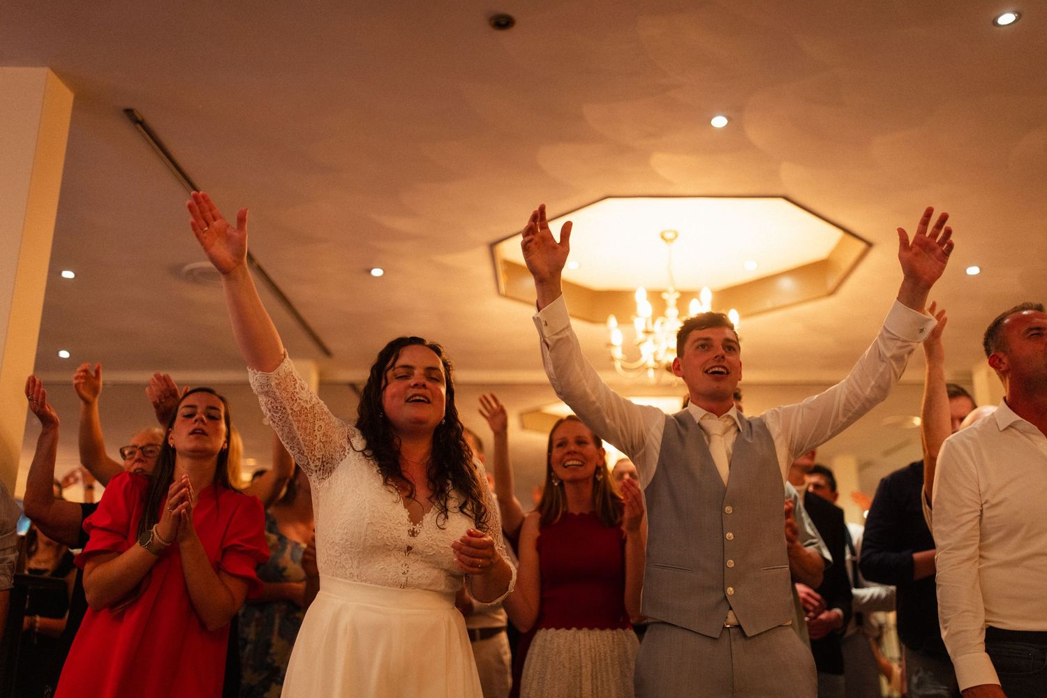 Newlyweds and wedding guests dancing with raised arms under warm reception venue lighting.