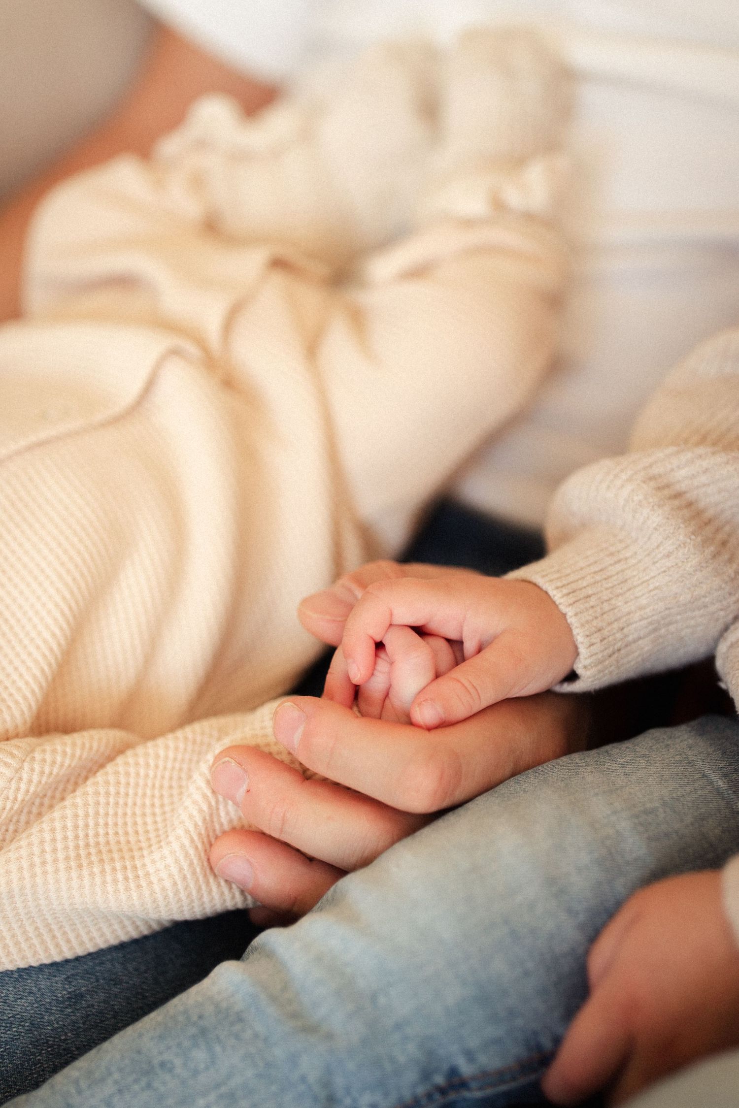 Close up of tiny baby hand holding parent's finger against cream colored fabric background.