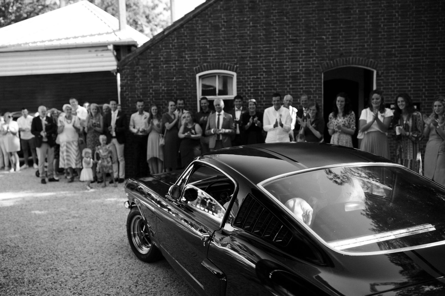 Vintage car parked outside brick building with crowd of people gathered for an event in black and white photo.
