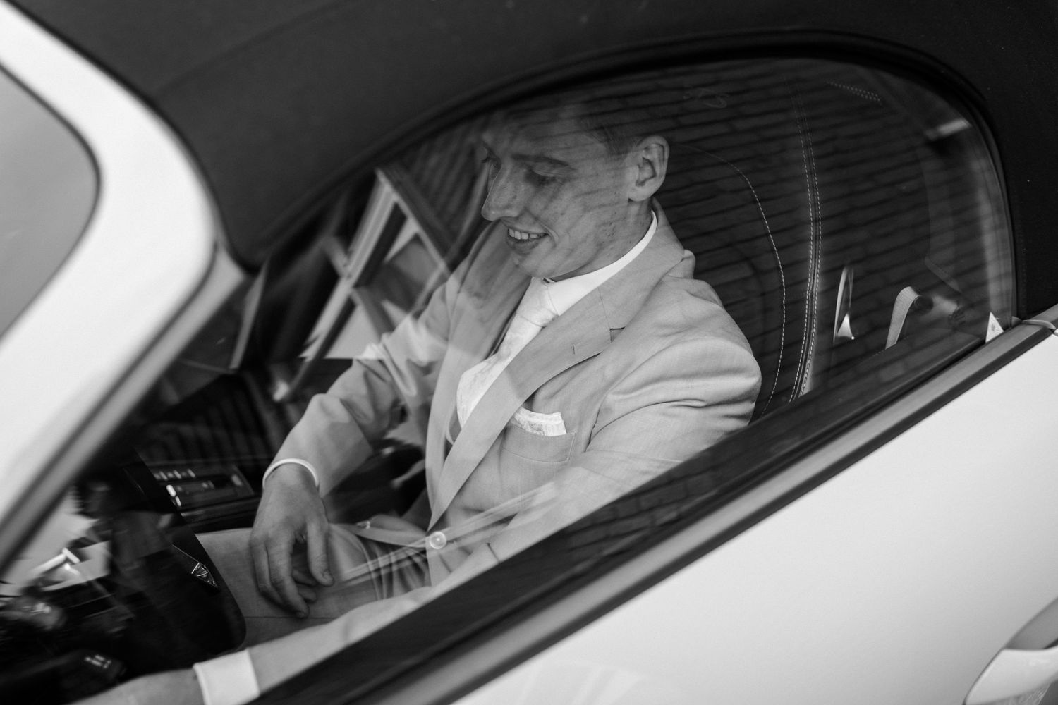 Black and white portrait of someone in formal attire sitting in driver's seat of car.