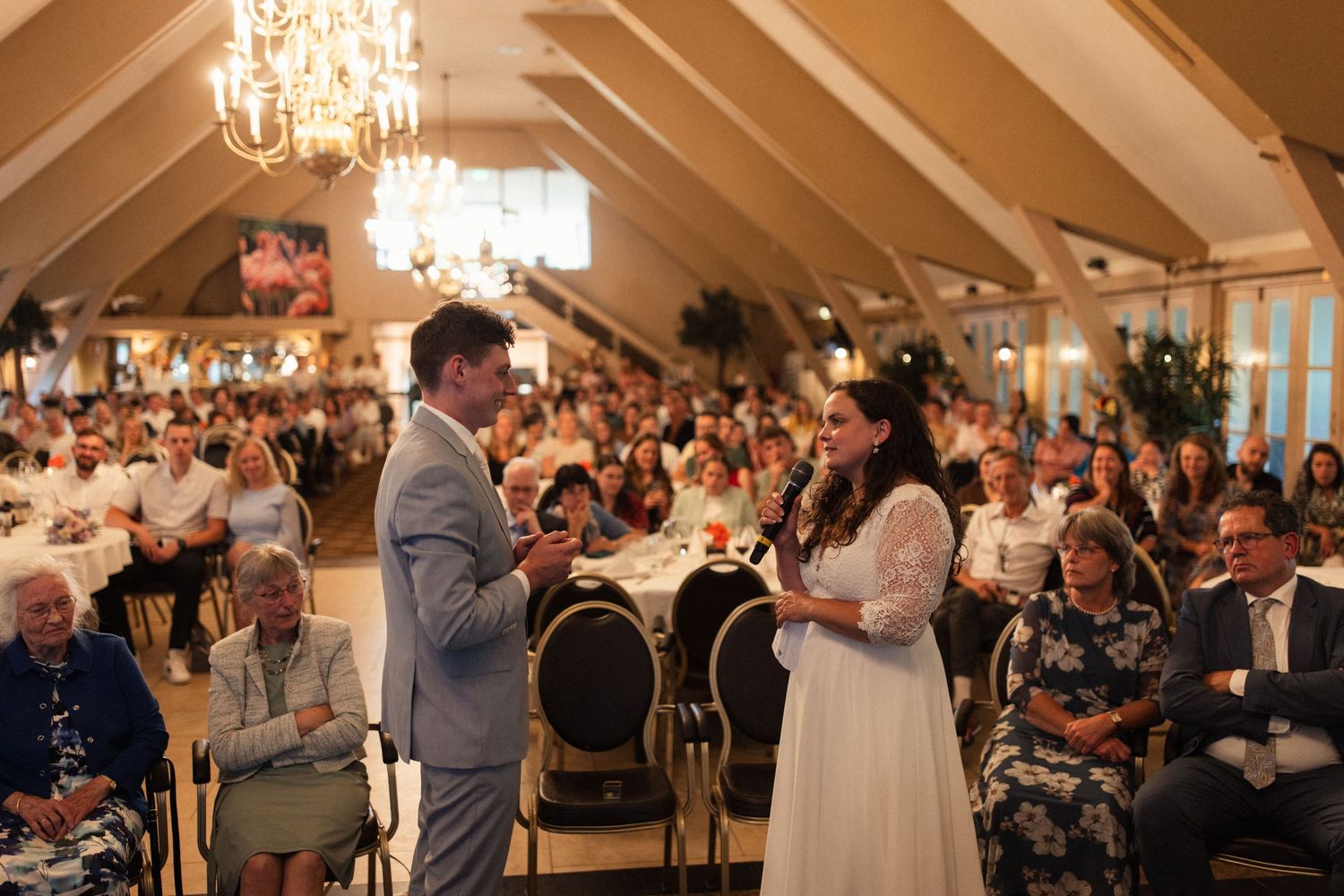 An elegant wedding ceremony takes place in a chandelier-lit venue with vaulted ceilings and a large audience.