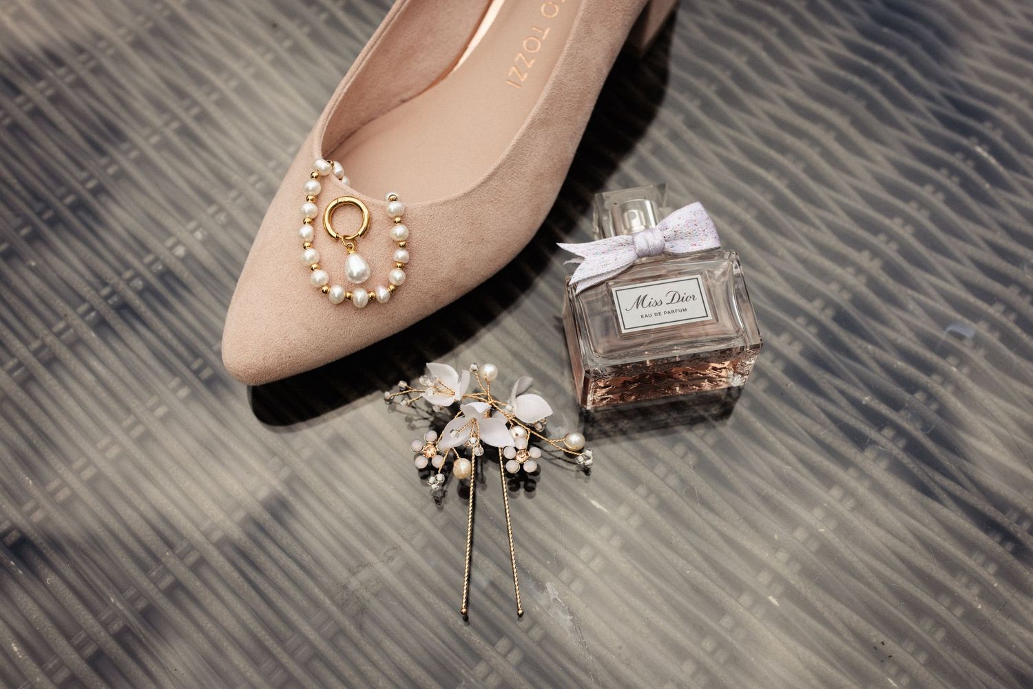 Wedding accessories arranged on gray carpet including nude ballet flat, perfume bottle and delicate pearl hair pin.