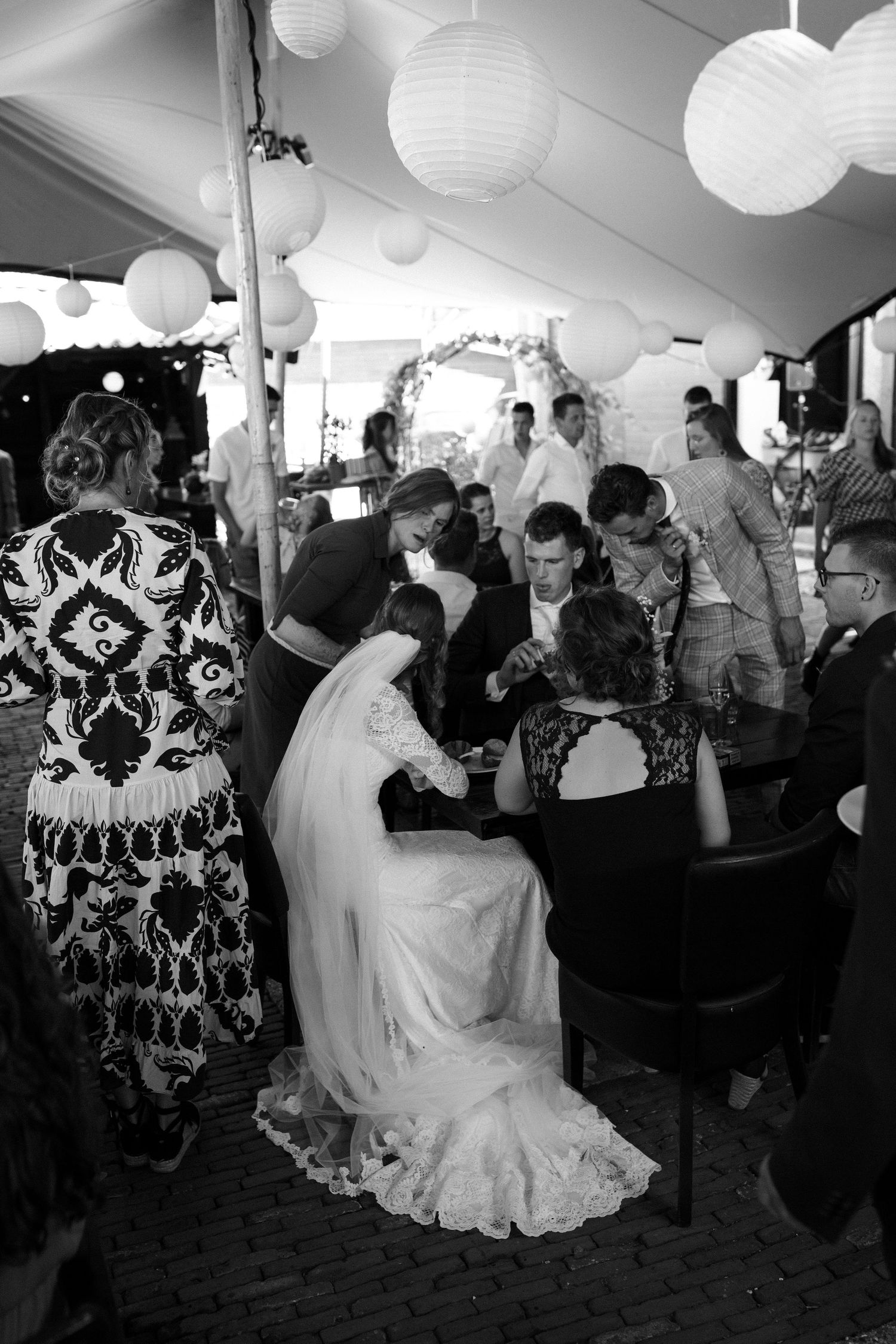 A black and white photograph of a wedding reception under a tent with white paper lanterns and guests gathered around.
