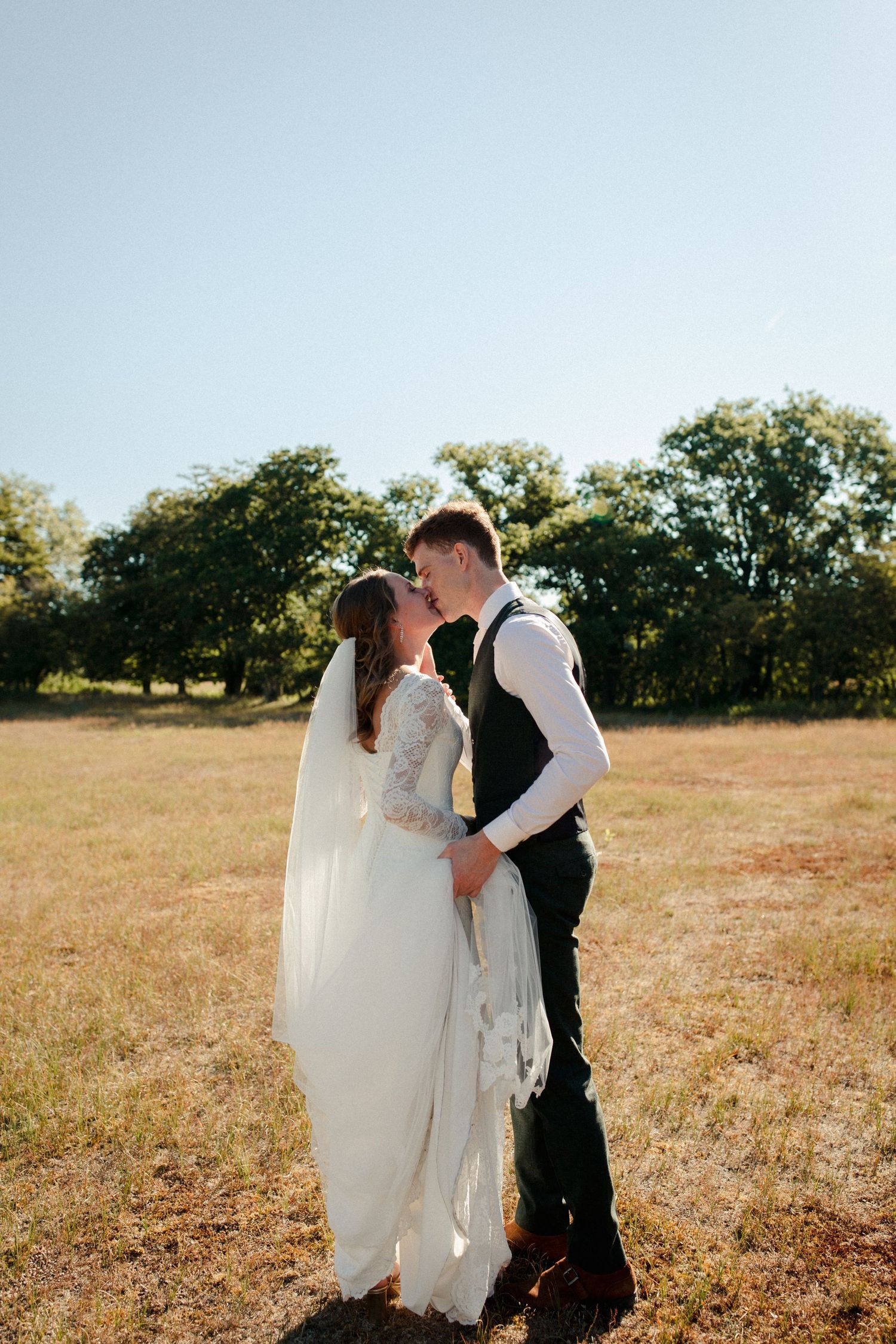 A couple shares a romantic kiss in a sun-drenched field with trees in the background.