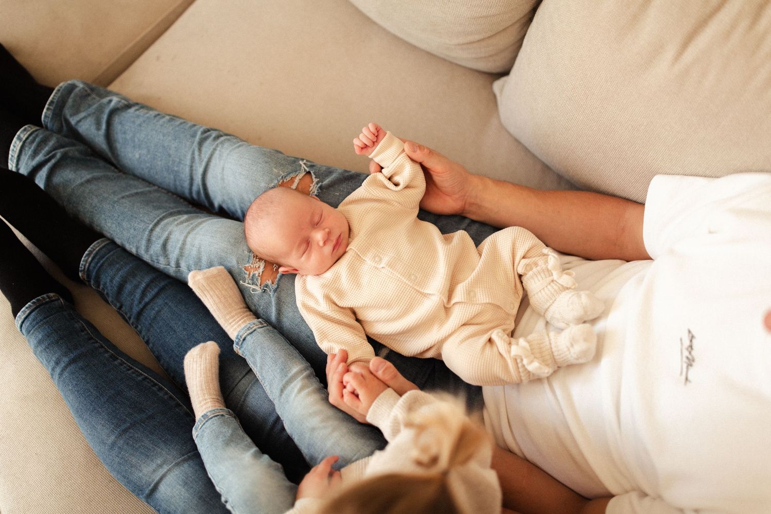 Sleeping newborn in cream outfit resting on parent's lap on beige couch.