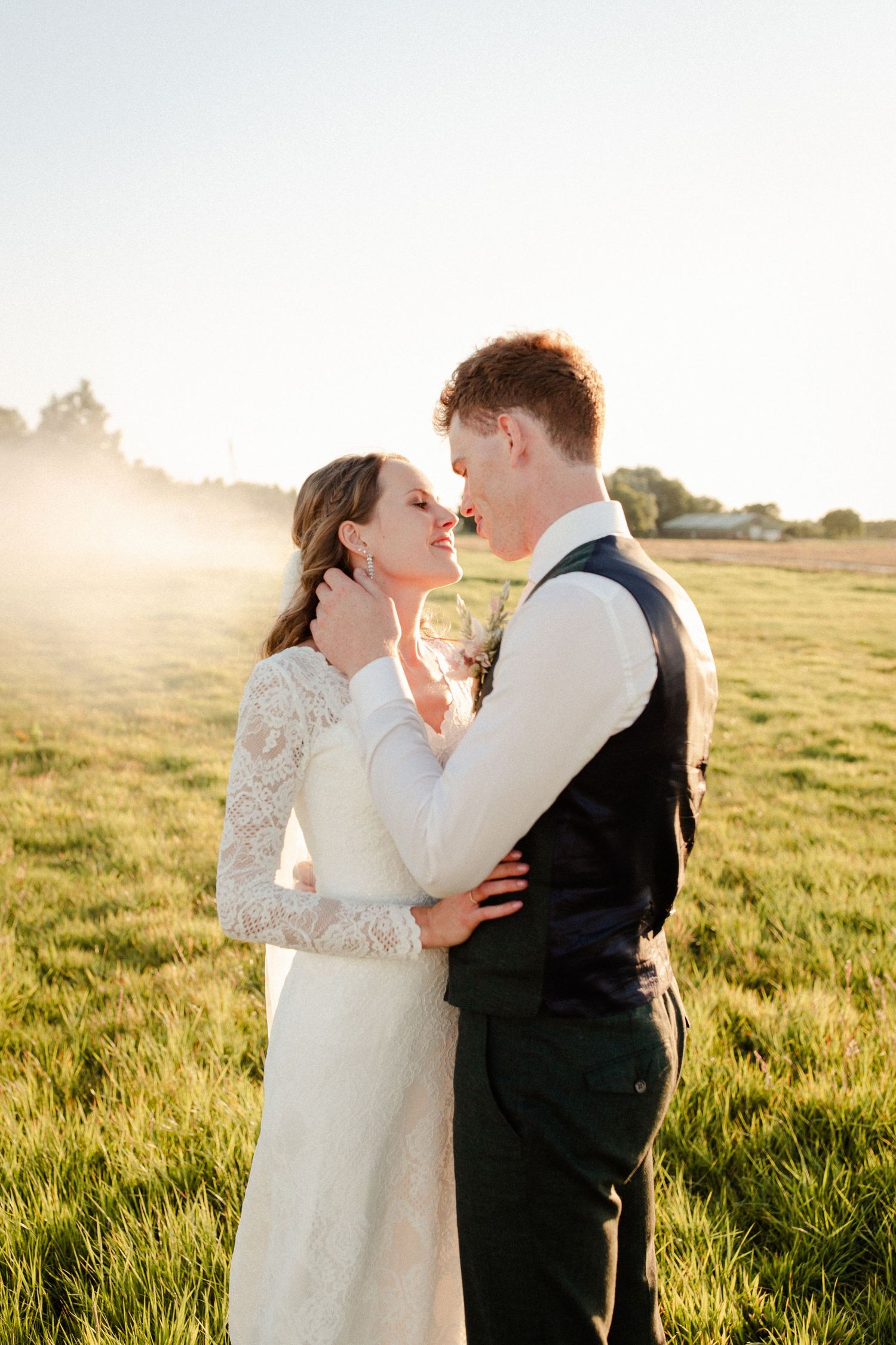 A romantic wedding couple embraces in a golden sunlit meadow during sunset, creating a dreamy outdoor portrait.