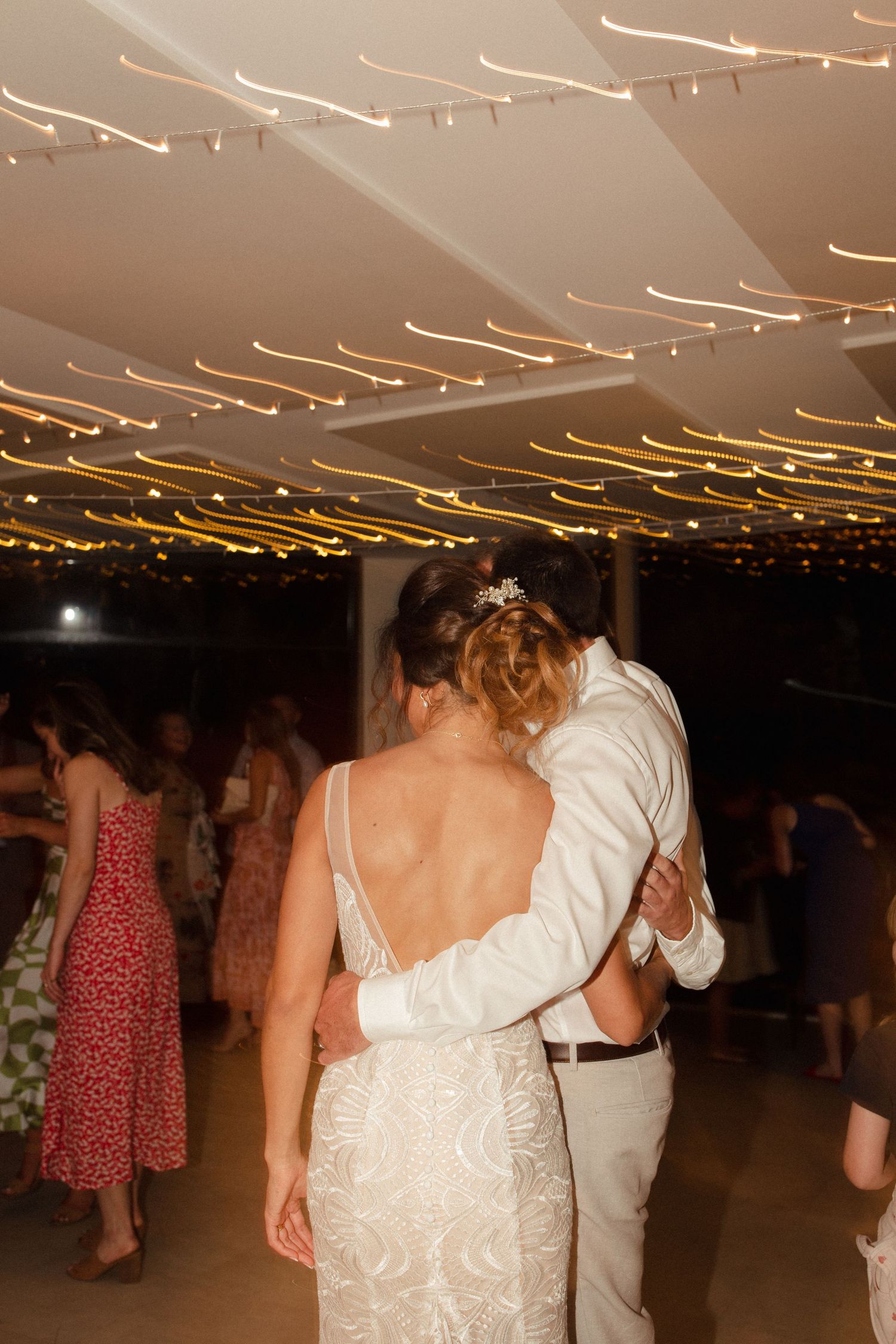 A couple embraces while dancing under golden string lights at an evening celebration.