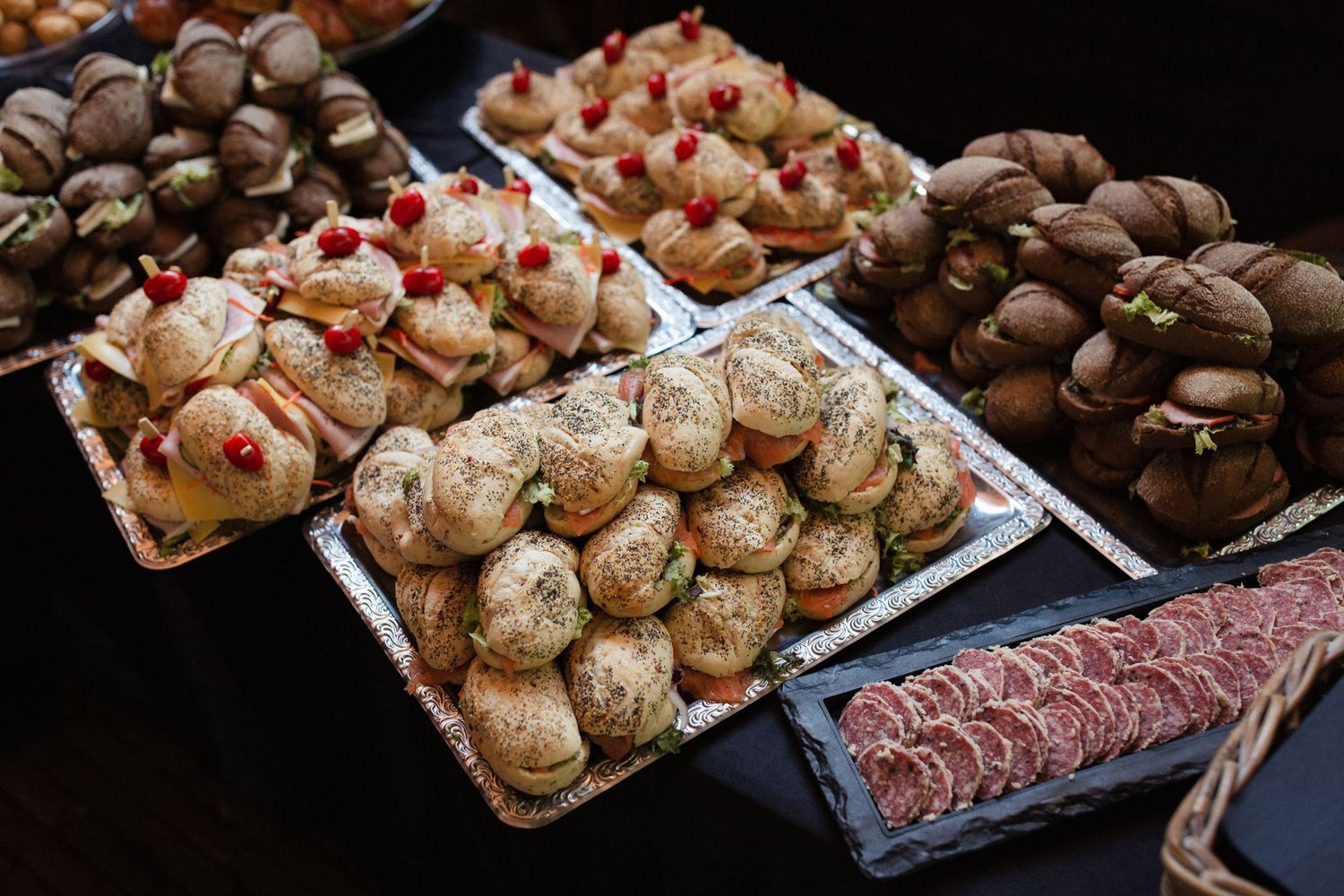 Platters of gourmet finger foods including mini sandwiches, charcuterie meats, and chocolate desserts arranged on a dark surface.