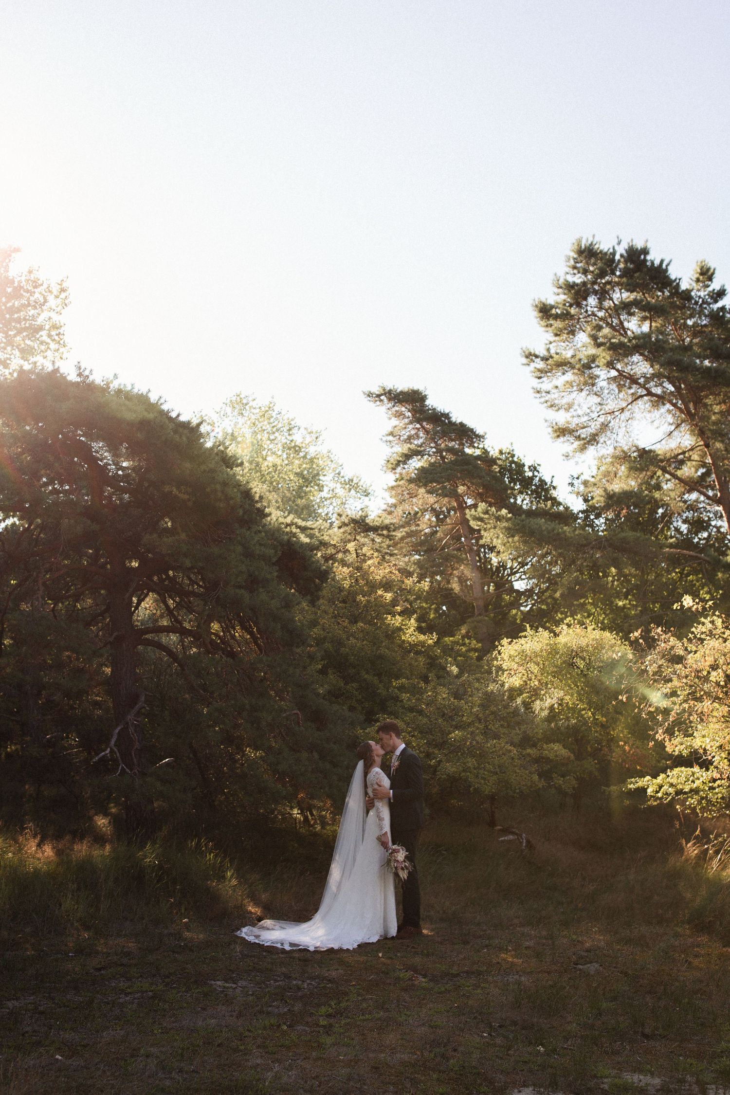 A couple in wedding attire stands together in a sunlit wooded area during golden hour.