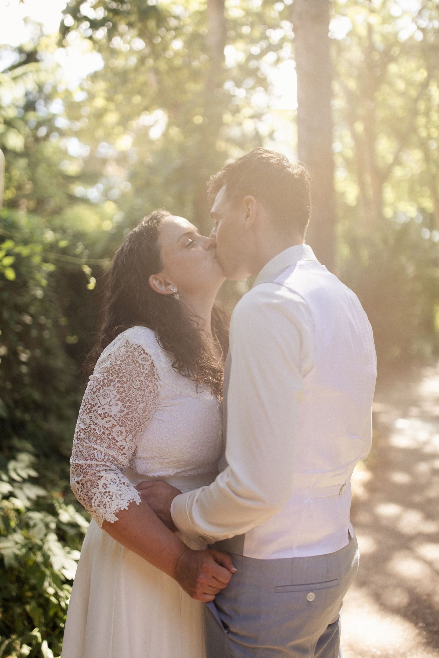 A couple shares a romantic kiss in a sunlit forest setting with golden hour lighting and the bride wearing a lace dress.